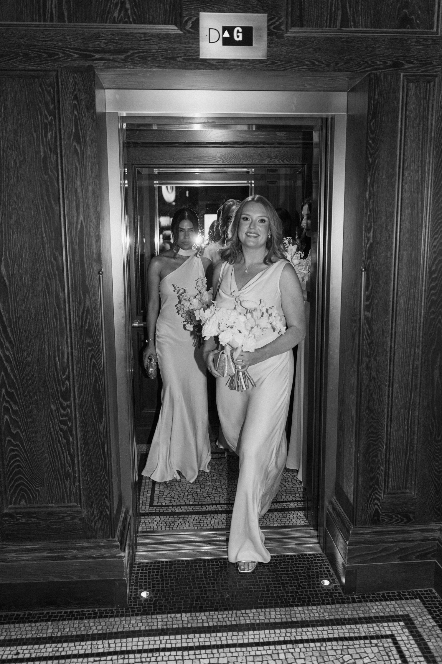Bridesmaid in elegant gown holding white bouquet stepping out of ornate elevator at wedding venue.