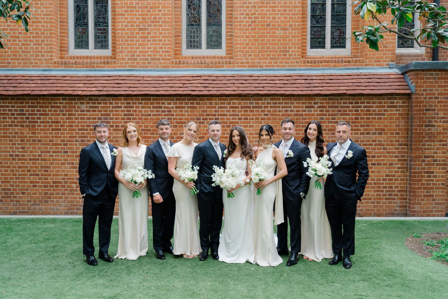 Wedding party poses together on grass in front of a red brick building with white bouquets.