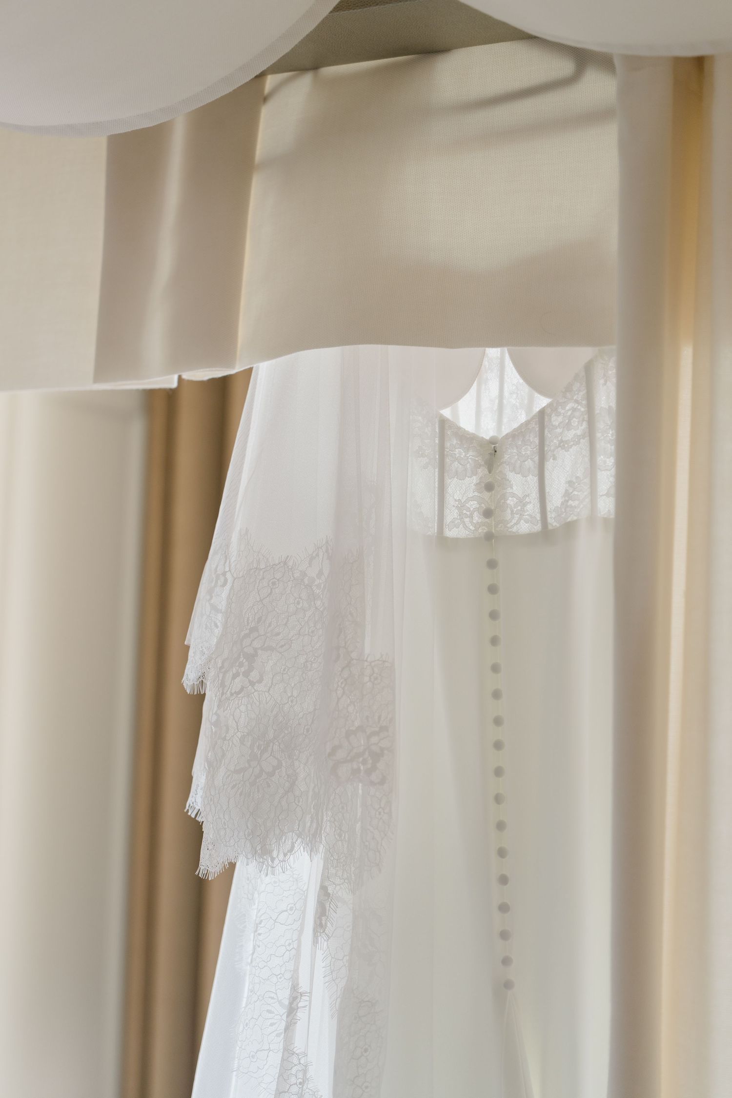 Elegant white wedding dress with lace veil hanging on a hanger against soft cream curtains in a bridal suite.