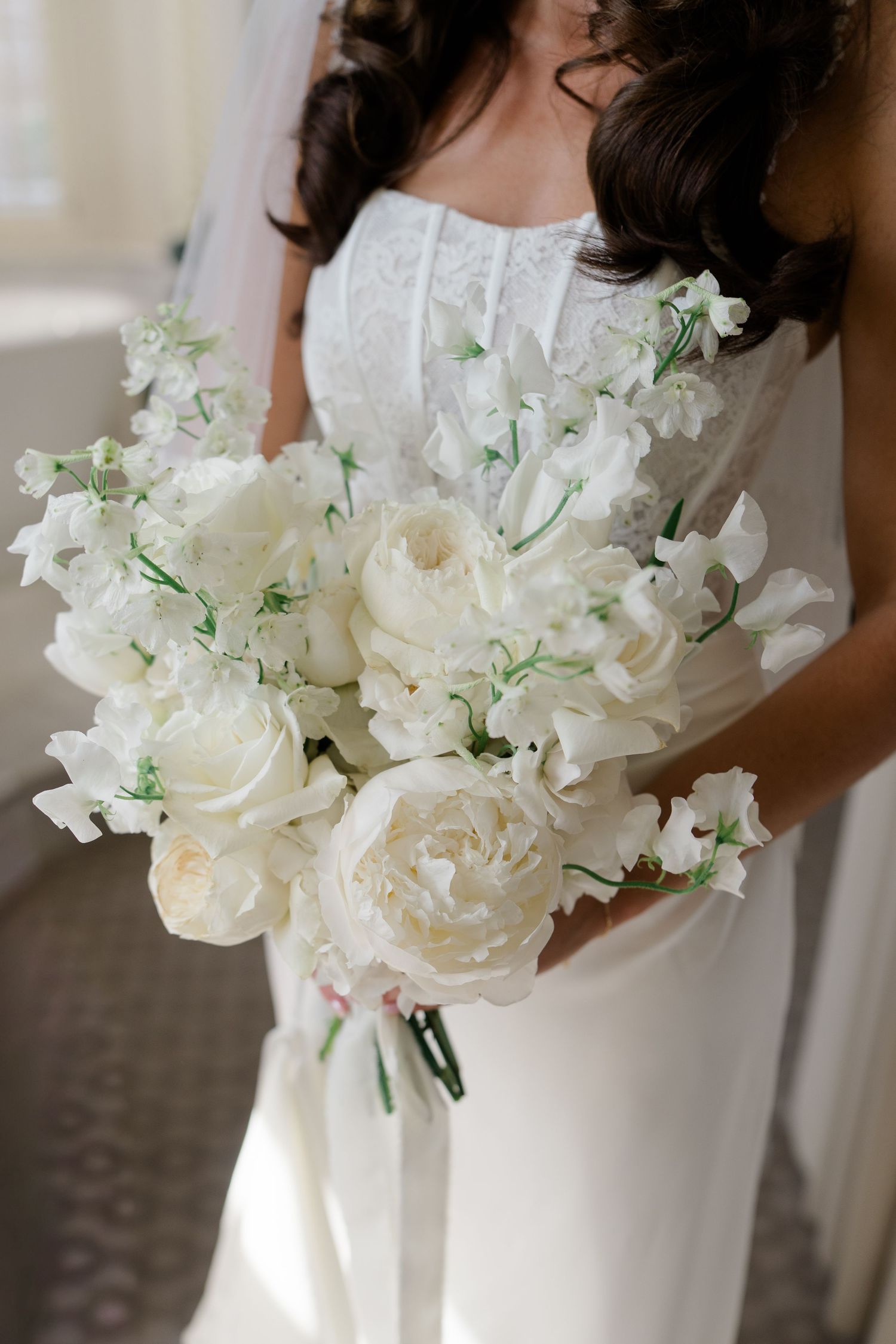 Bride in white lace gown holding elegant all-white bouquet of peonies and sweet peas with green stems.