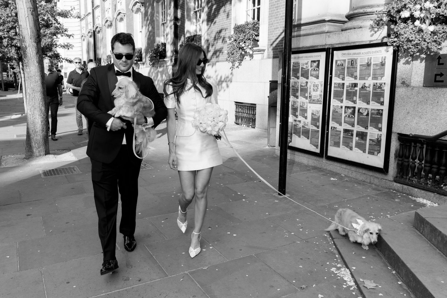 Black and white photo of an elegant couple walking dogs on a city sidewalk, woman in mini dress, man in suit carrying a small dog.