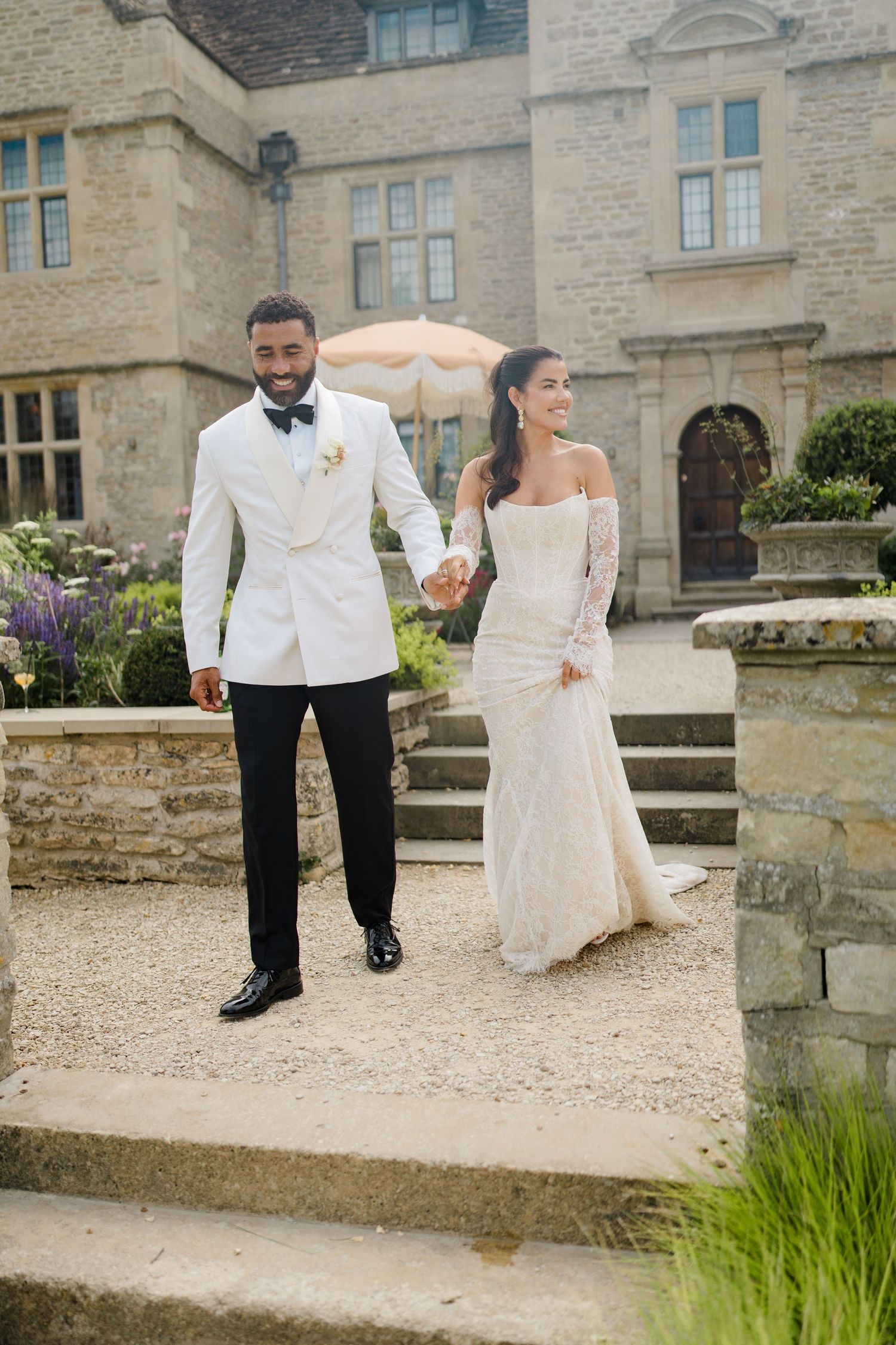 A joyful bride and groom in elegant attire hold hands while walking through the grounds of a stone manor house.
