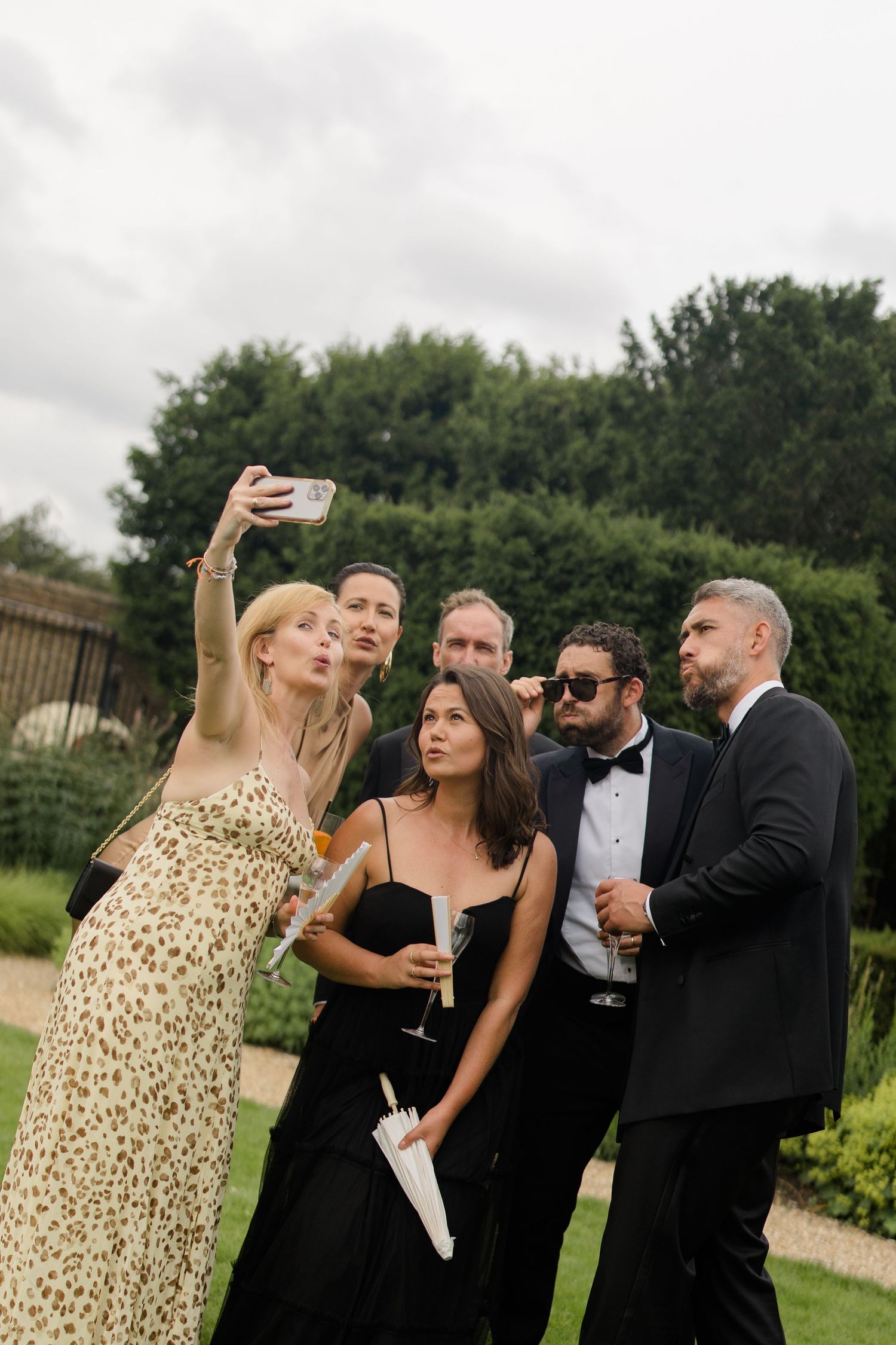 Wedding guests in formal attire taking a group selfie in lush garden at outdoor reception.