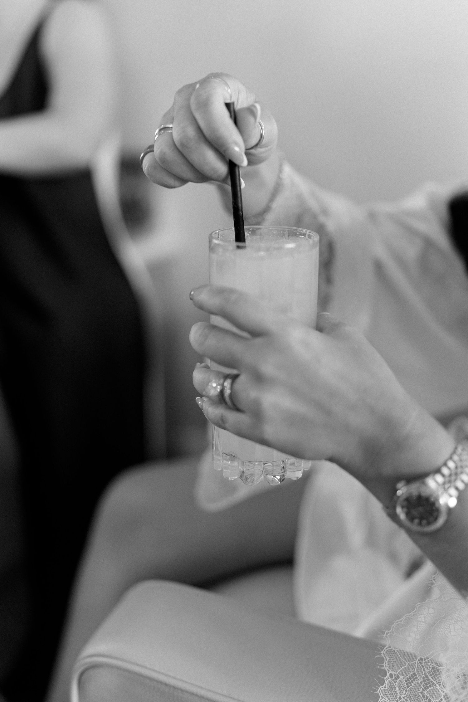 Black and white close-up of a bride in a white robe stirring a drink with a straw, wearing a watch.