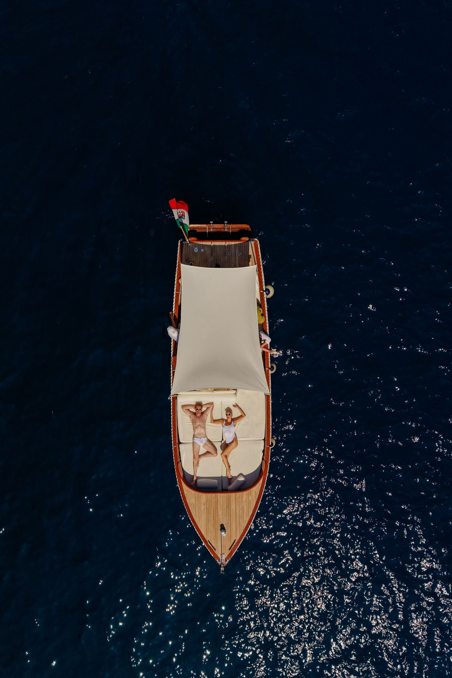 Aerial view of two people relaxing on the deck of a wooden boat floating on deep blue ocean water.