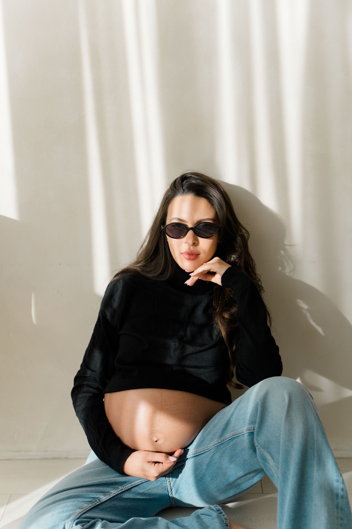 Pregnant woman in black crop top, jeans, and sunglasses sitting against white wall in natural light.