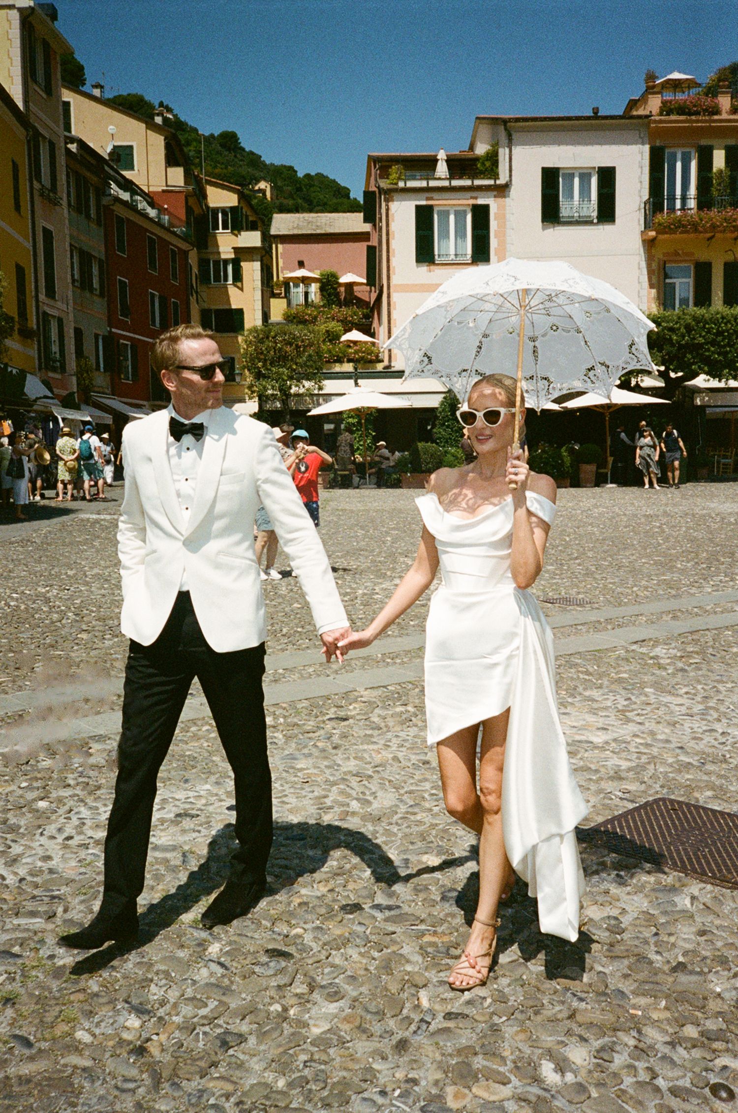 Bride and groom holding hands in a sunny Italian piazza, she carries a white parasol wearing a sleek wedding dress.