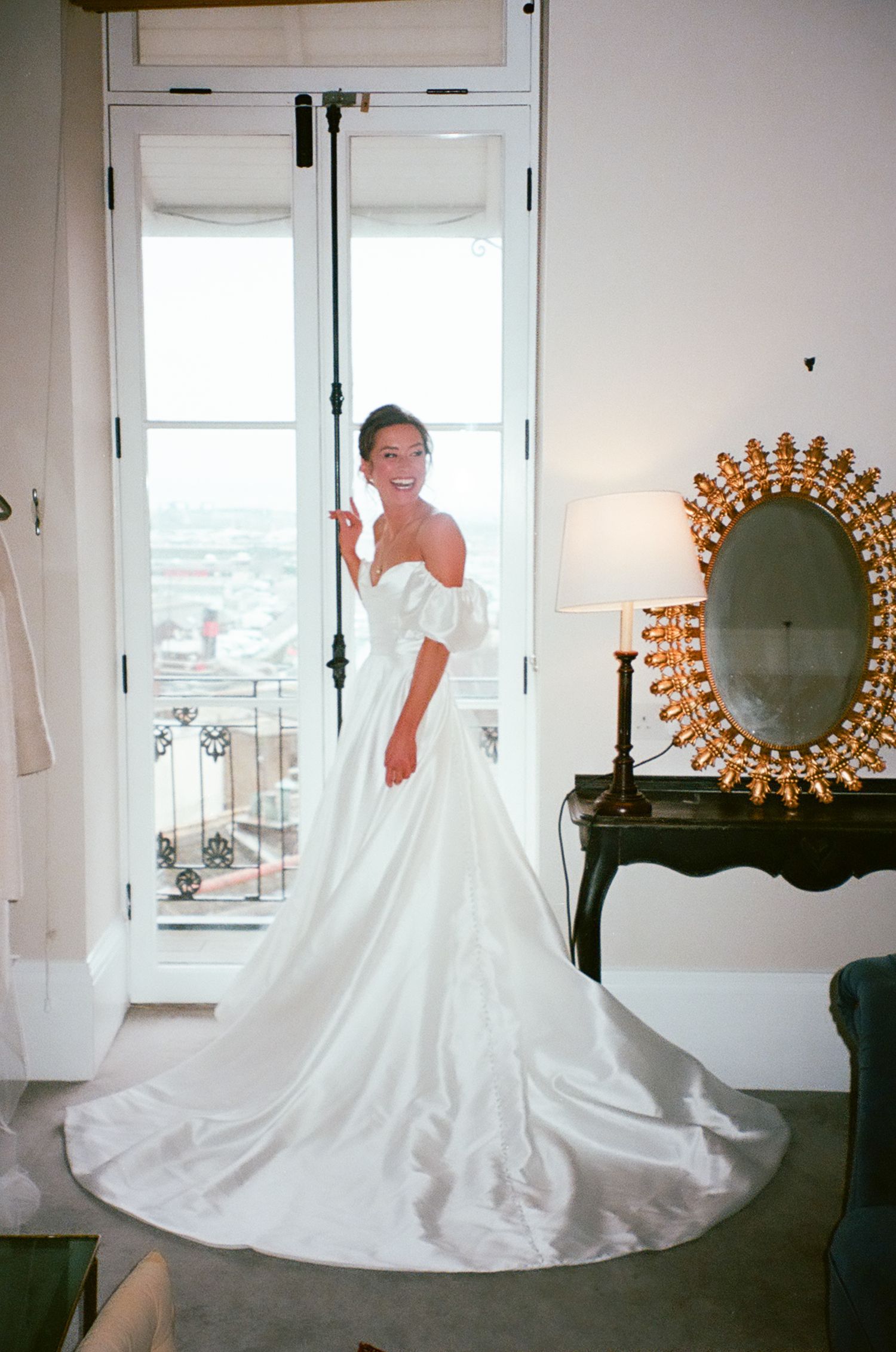 An elegant off-shoulder white wedding gown with a flowing train is showcased in a bright room with a decorative sunburst mirror.