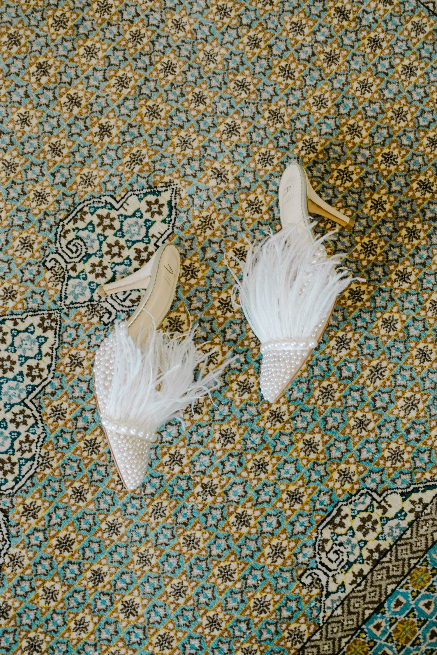 Elegant white feathered bridal shoes with pointed toes displayed on an ornate blue and gold patterned background.
