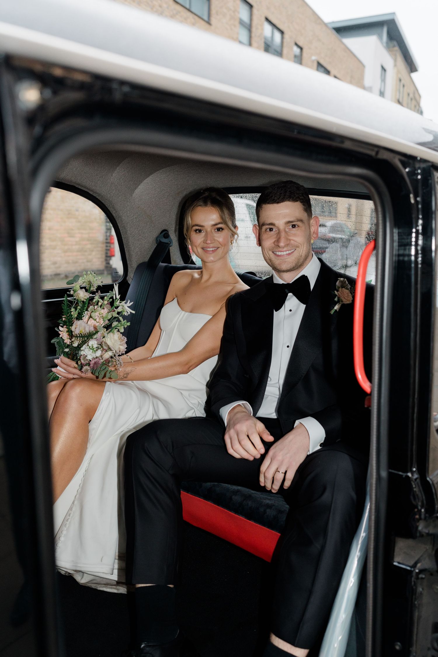 Wedding couple smiles and embraces in the back seat of a vintage black car during their celebration.
