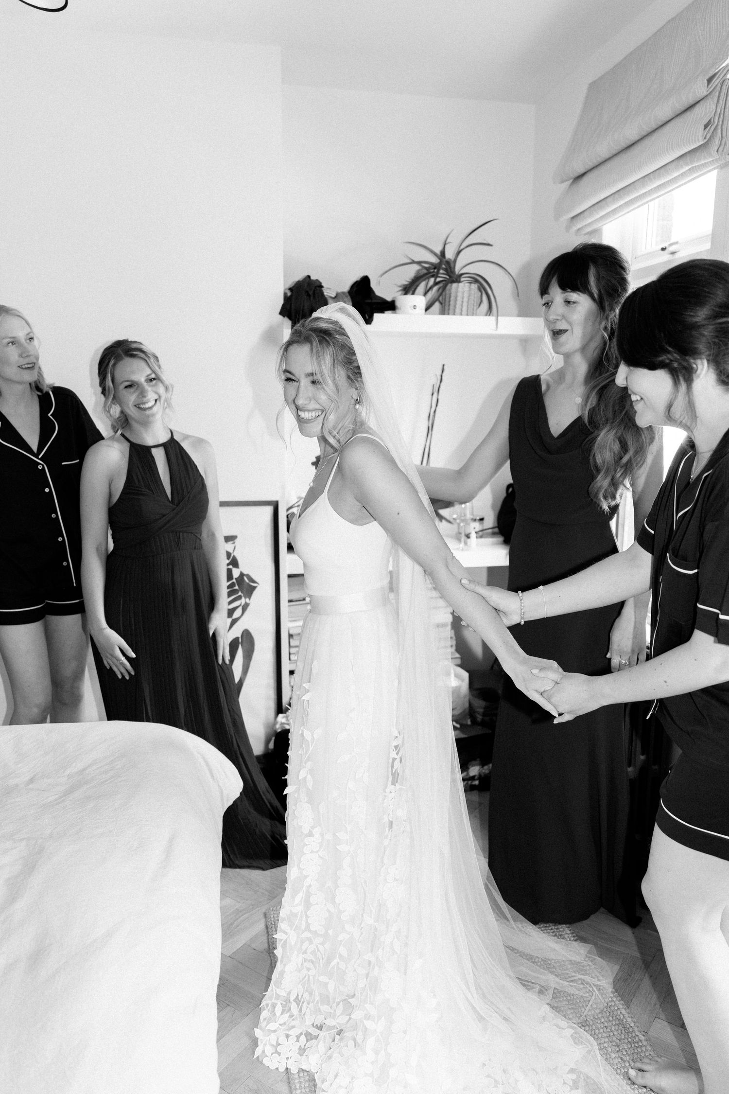 A bride in a lace wedding gown gets ready with her bridesmaids wearing black dresses.