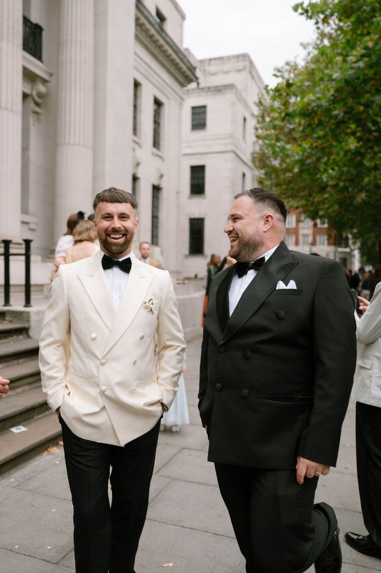 Wedding party members in black and white tuxedos share laughs outside a columned building.