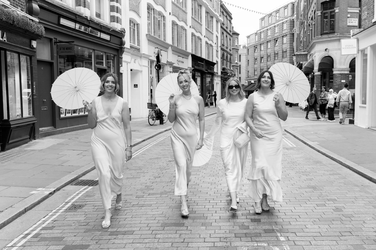 Four people in white dresses walk down cobblestone street carrying parasols in black and white photo.