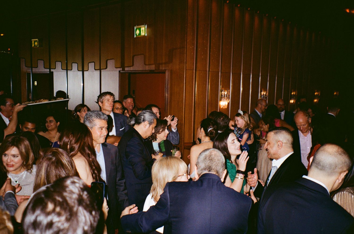 A crowded wedding reception shows guests mingling and dancing in a wood-paneled venue.