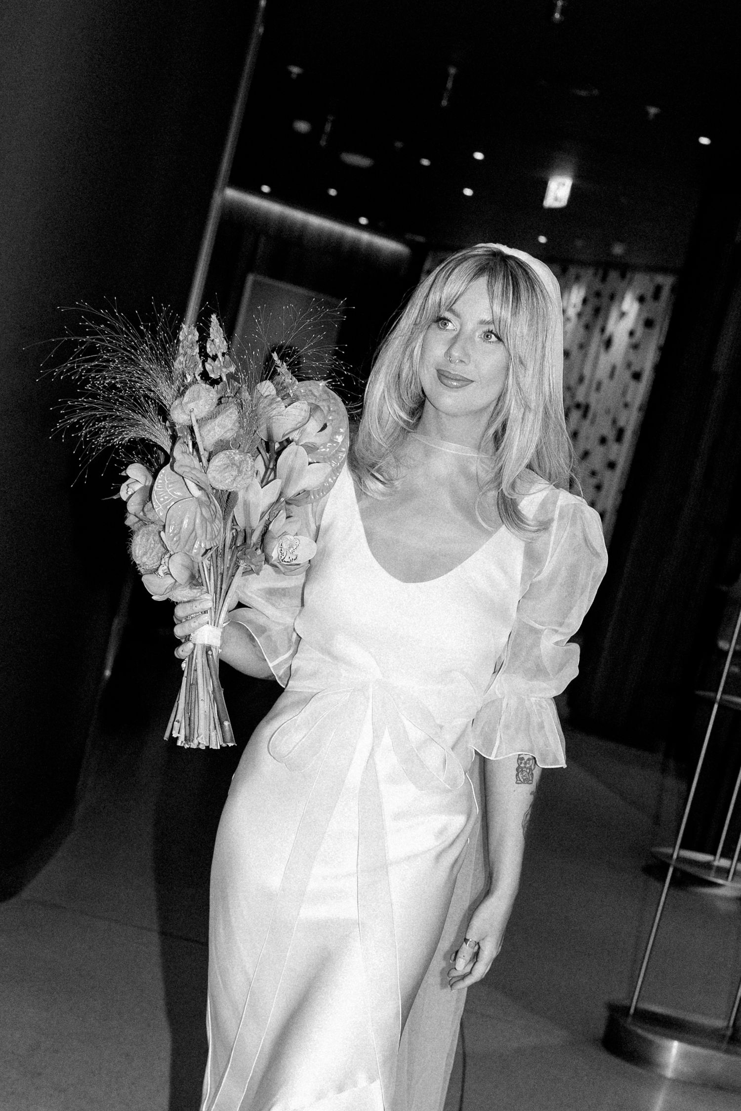 Black and white photo of elegant white wedding dress with bouquet in formal setting.