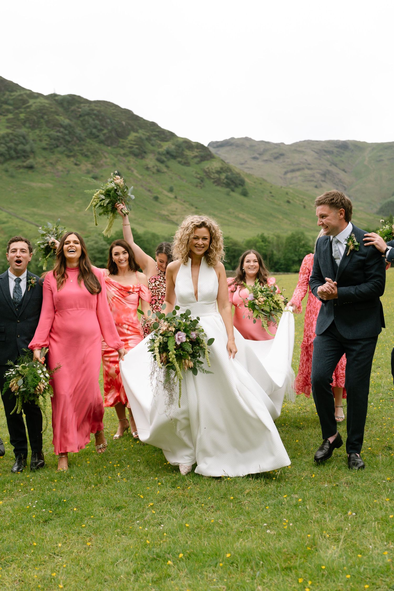 Wedding party in coral dresses and dark suits poses on grassy hillside with mountains in background.