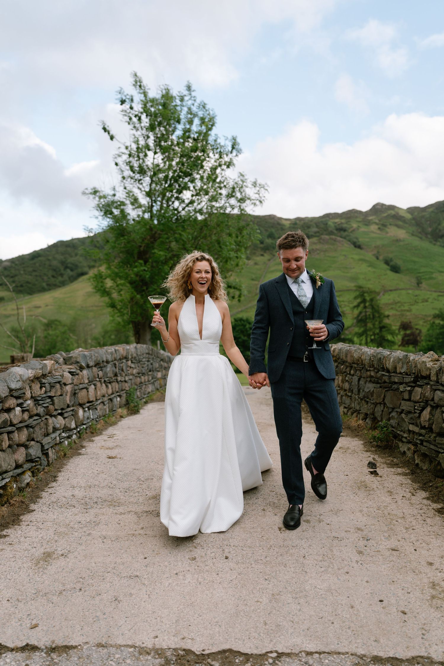 A couple walks along a stone-walled path with rolling hills in the background.