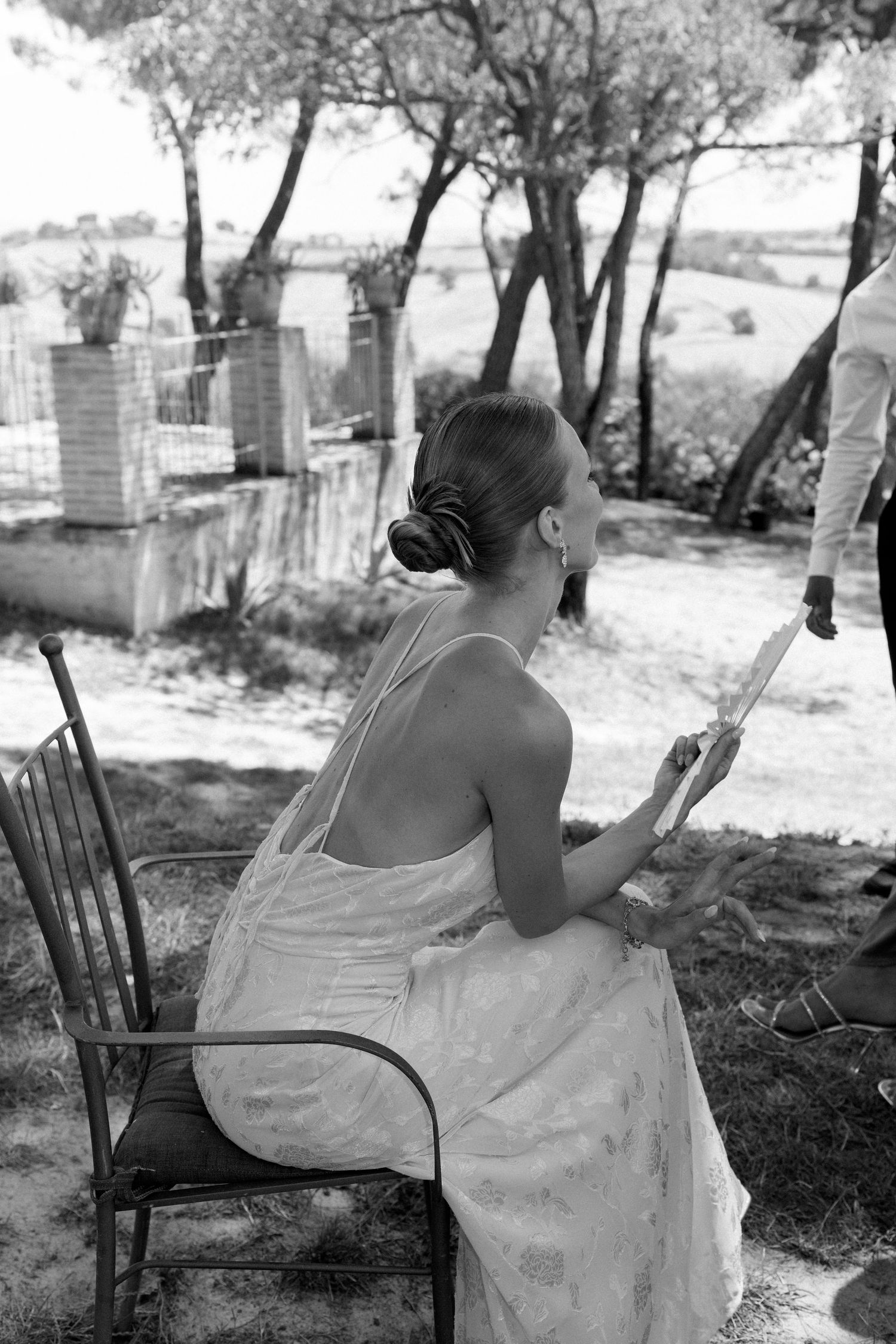 A person in a white dress sits on a wooden chair in a garden setting.
