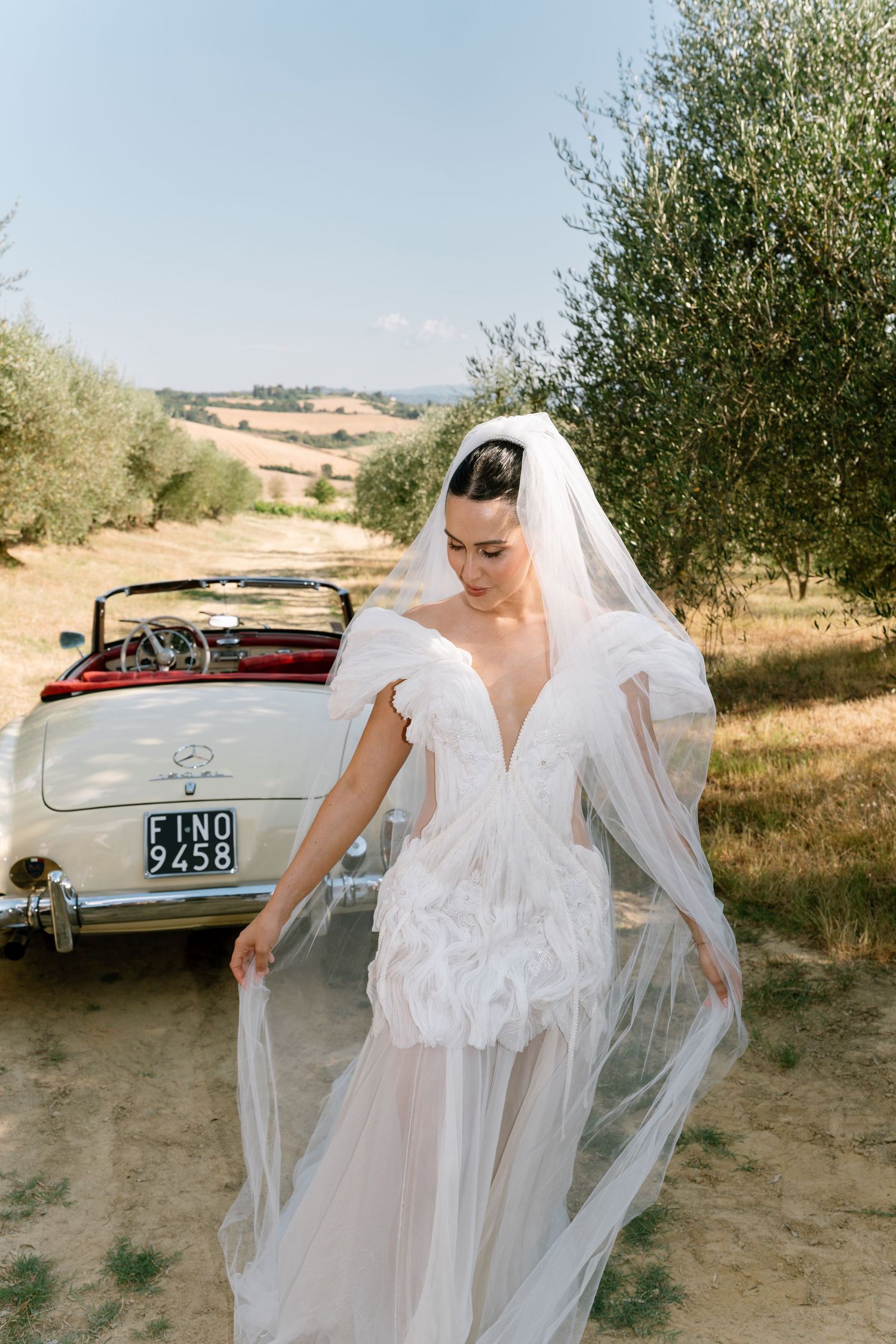 A flowing white wedding dress with delicate lace details captured in a rustic outdoor setting with a vintage car.