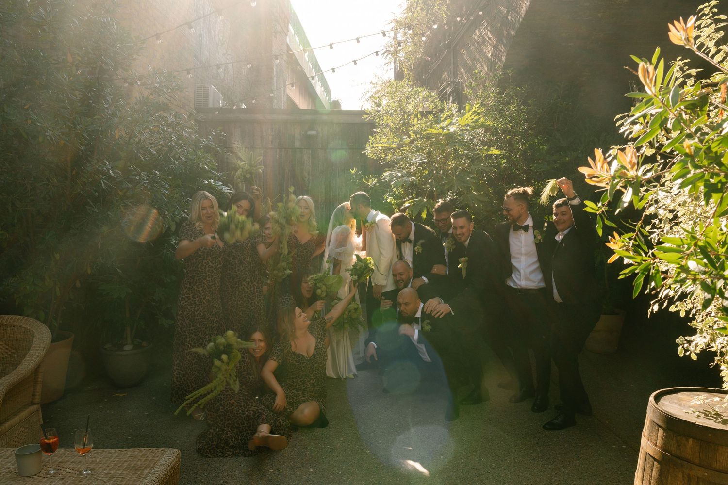 Wedding ceremony guests gather in an outdoor garden setting with golden sunlight filtering through trees.