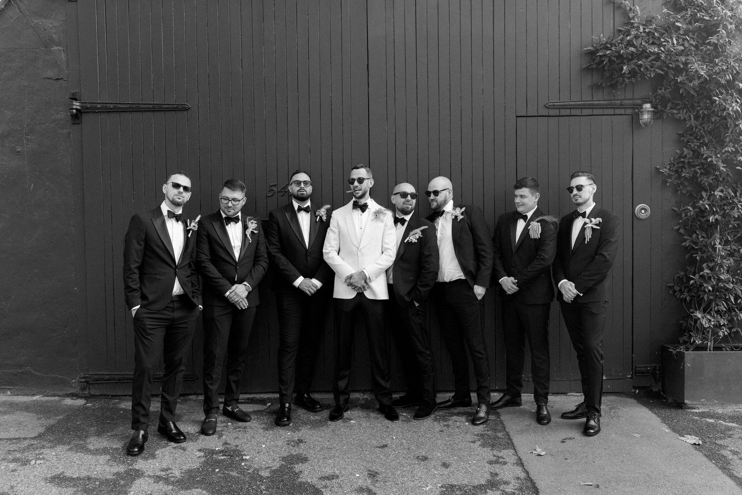 Wedding party in formal attire poses against dark wooden wall in outdoor setting.