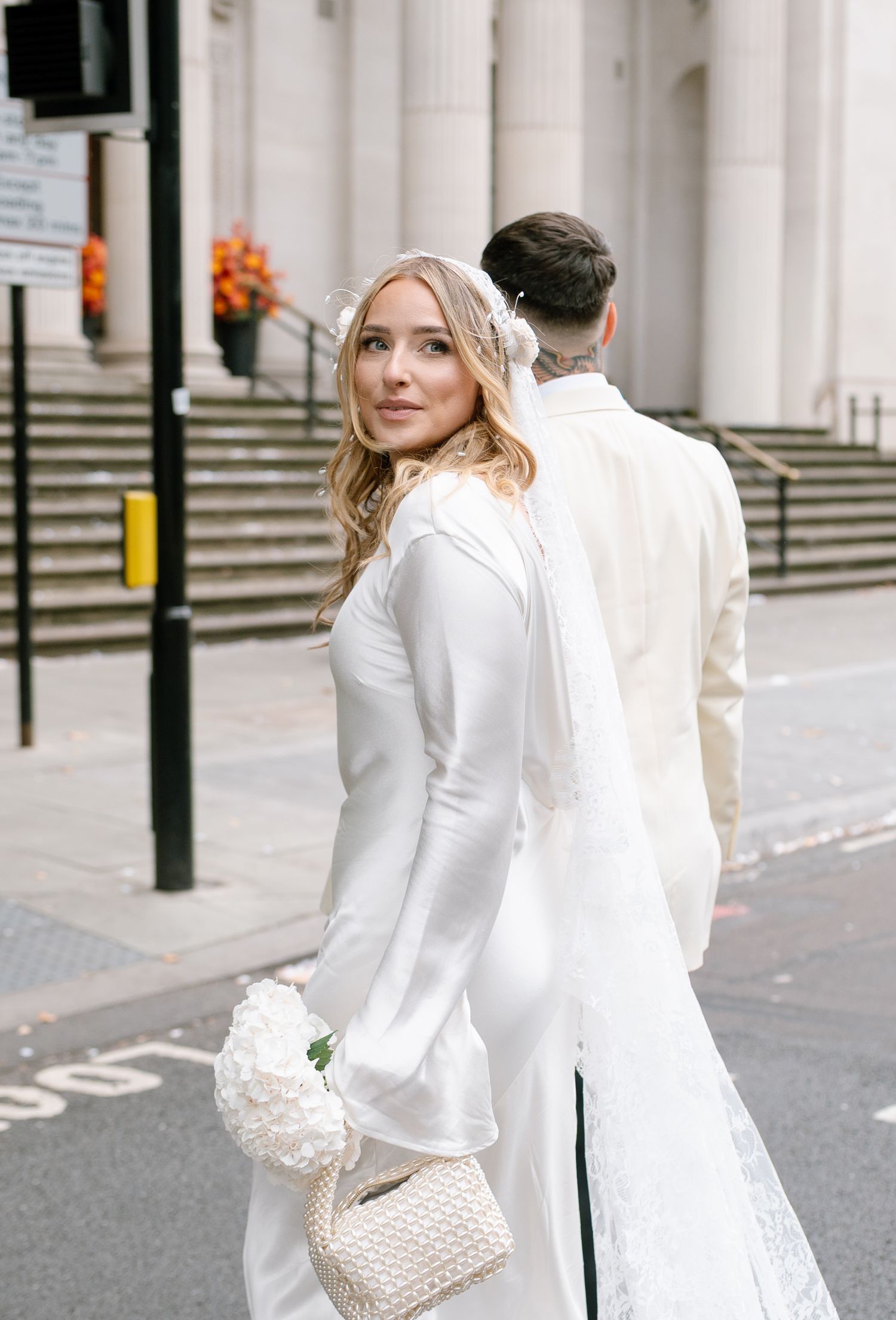 Wedding couple in all white attire walking near classical building columns.