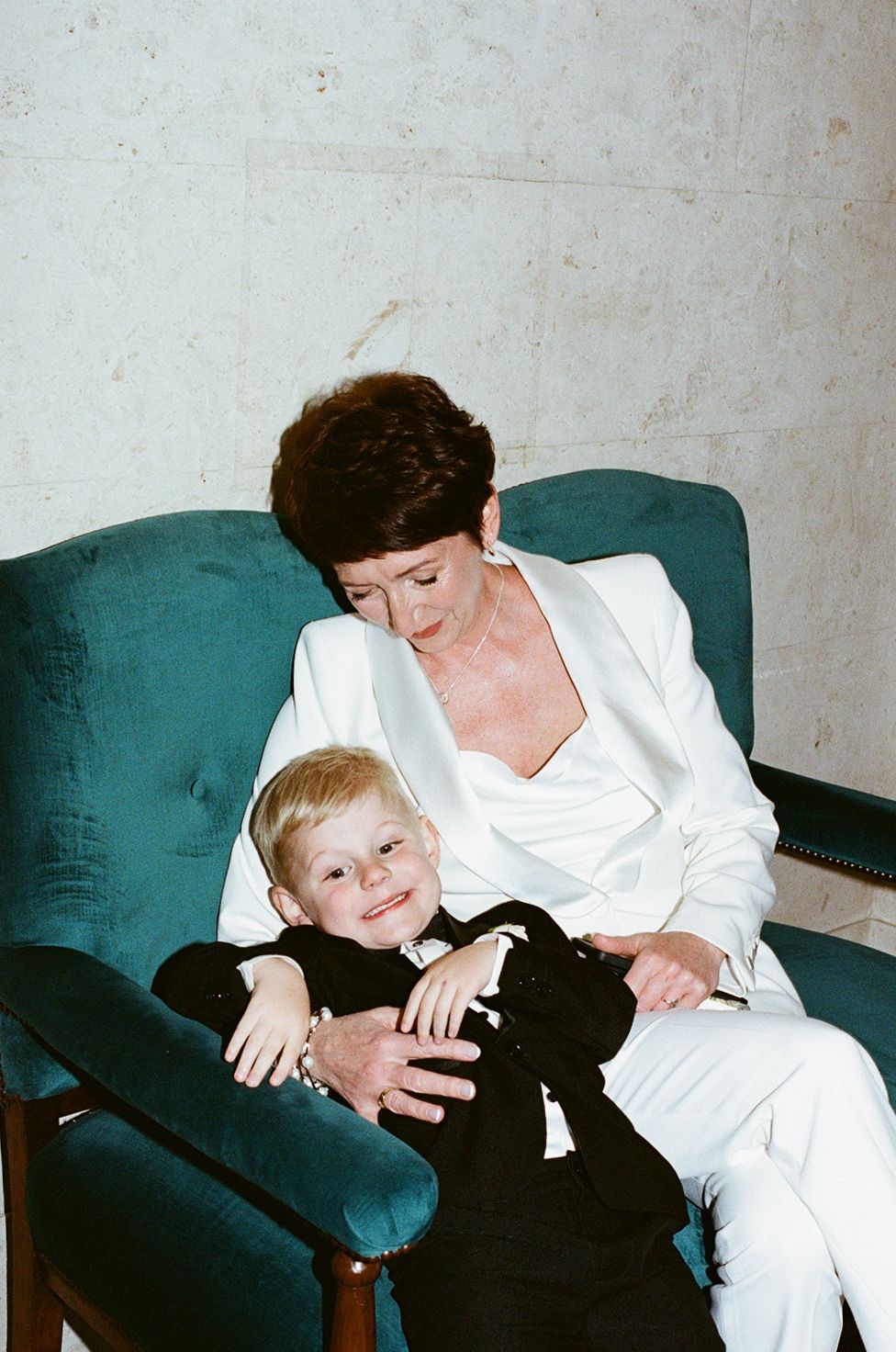 Woman in white suit holding a smiling blonde toddler in black outfit on a teal armchair.