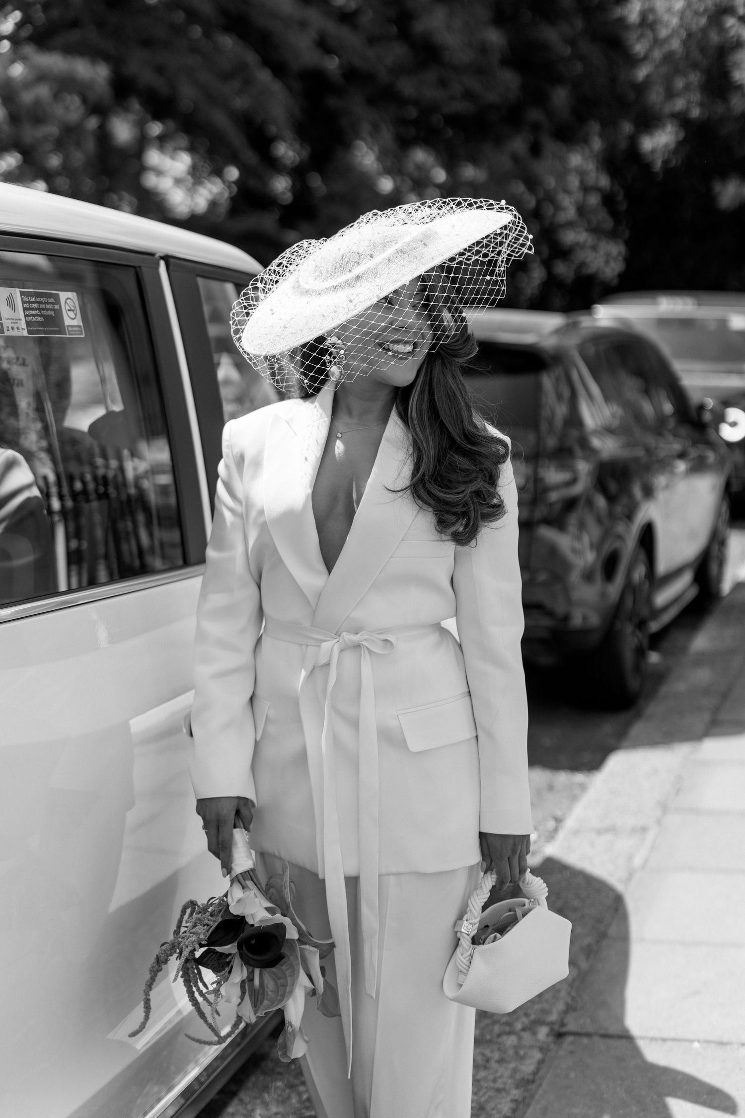 Elegant bride in white pantsuit and birdcage veil hat standing beside a car on her wedding day.
