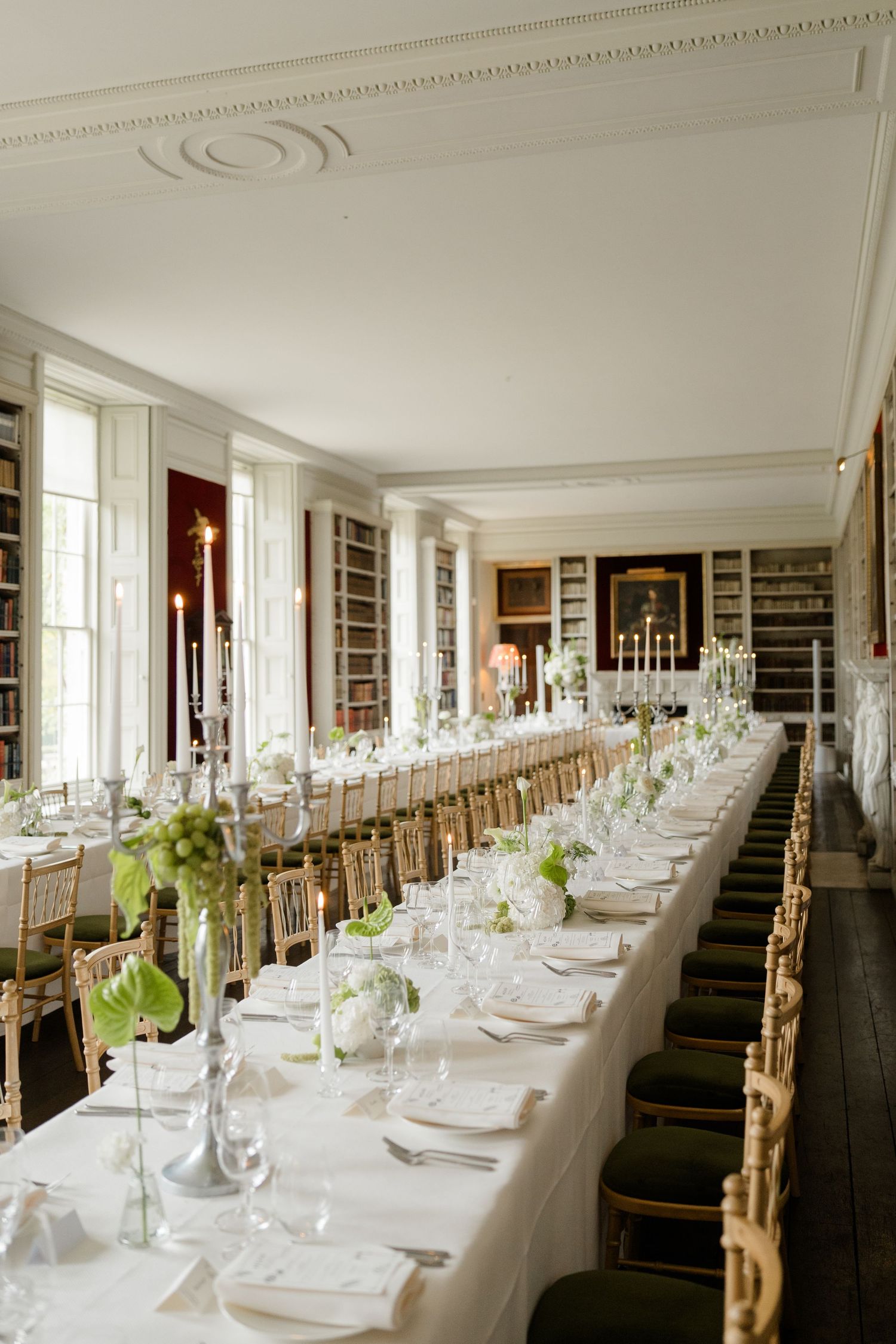 Elegant wedding reception in a library with long white tables, gold chairs, and green floral centerpieces.