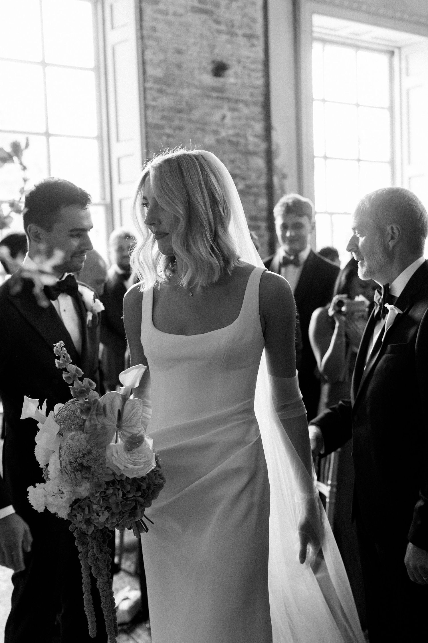 Bride in sleek white square-neck gown walking down aisle with bouquet at wedding ceremony.