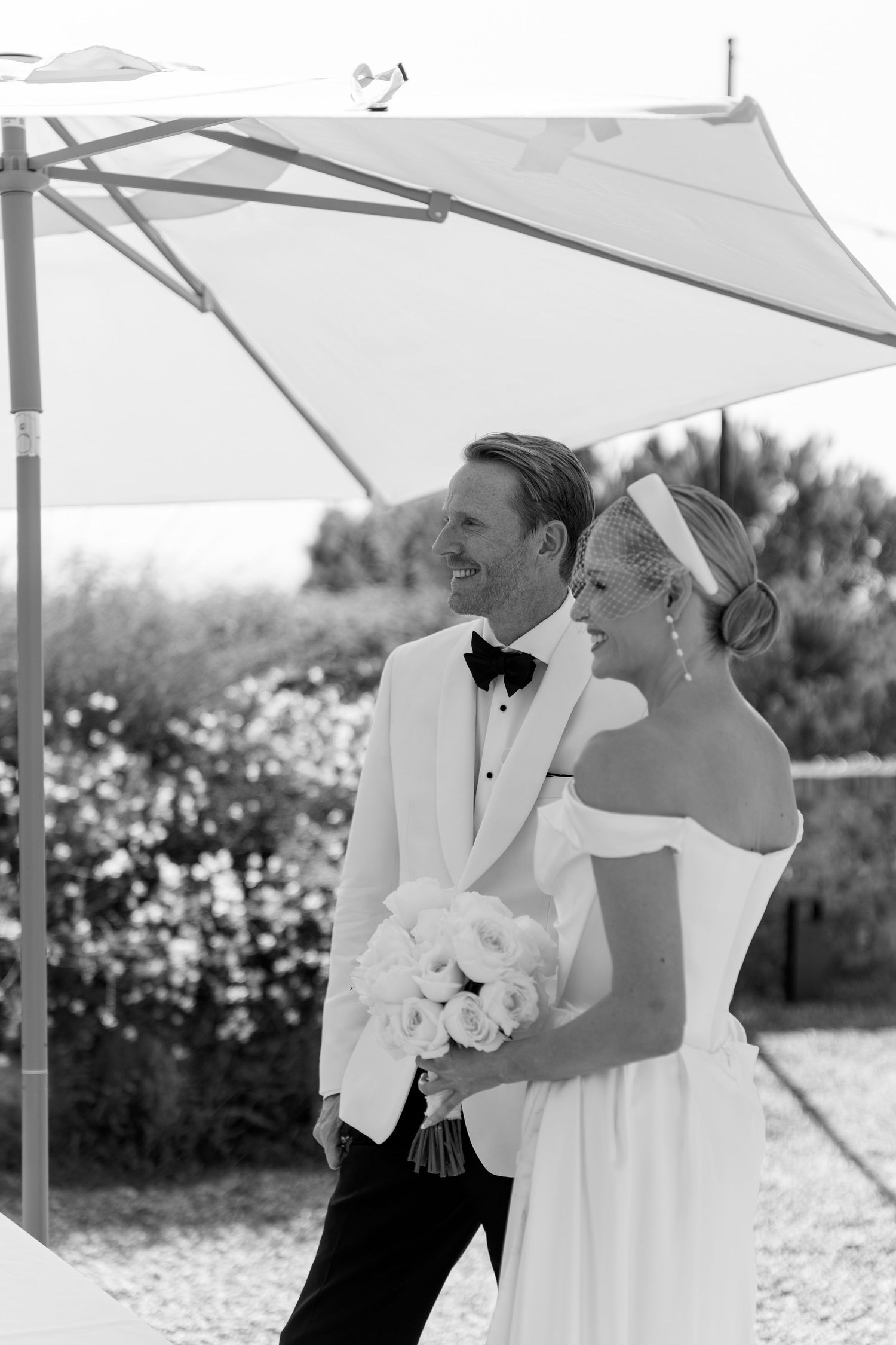 Smiling bride and groom stand under umbrella outdoors, bride holds white rose bouquet.