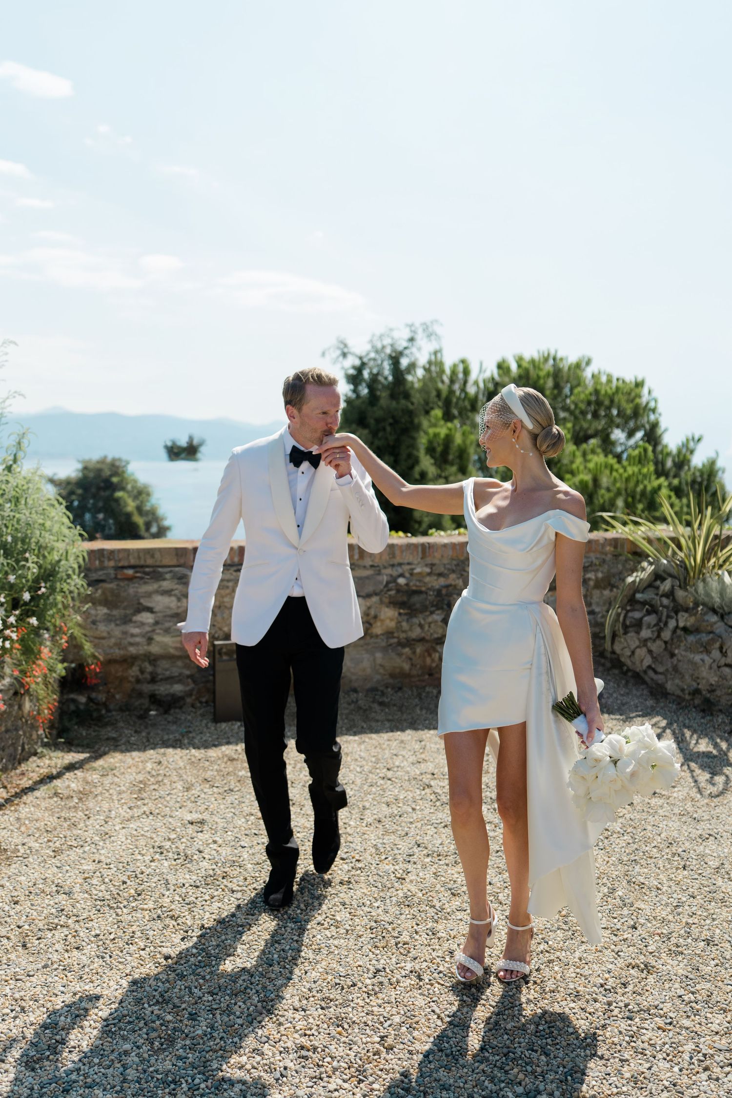 Bride in a short white off-shoulder dress and groom in a white tuxedo walking together on a sun-drenched terrace.
