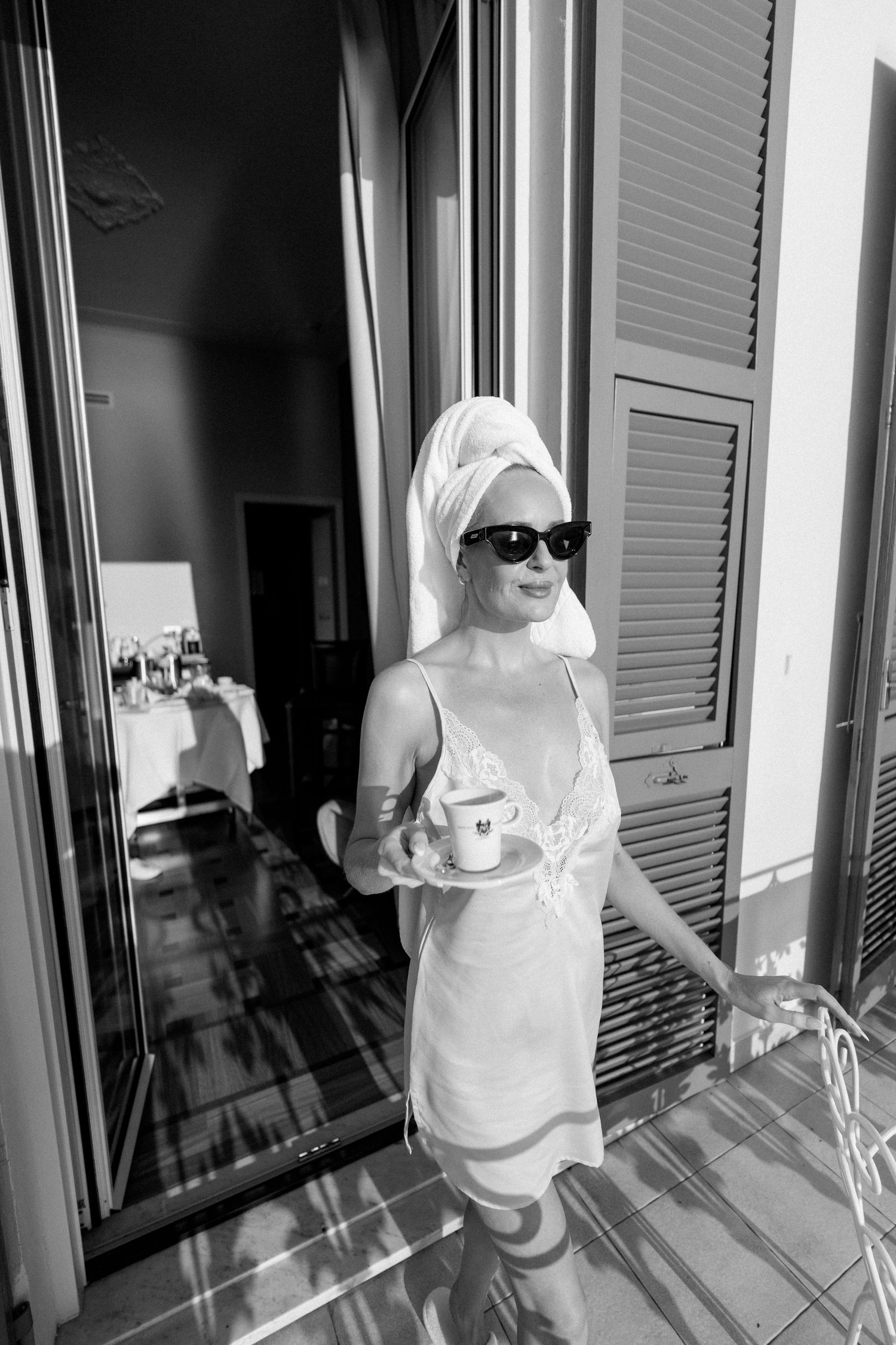 Elegant woman in white slip dress and towel turban holding coffee cup on sunny hotel balcony.