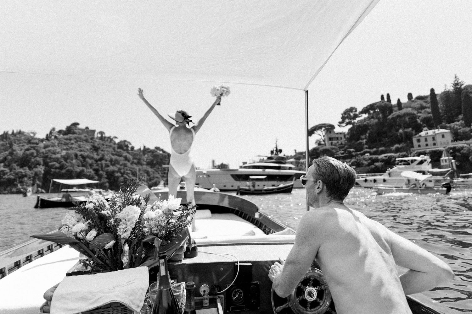 Jubilant bride raising bouquet triumphantly on boat bow in scenic Italian harbor setting.