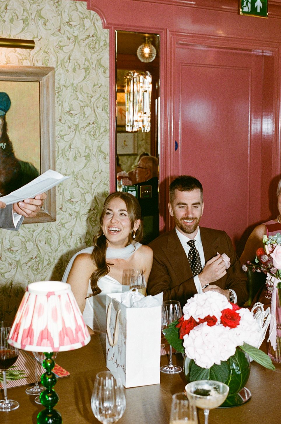 Bride and groom laugh joyfully at wedding reception table with flowers and wine glasses.