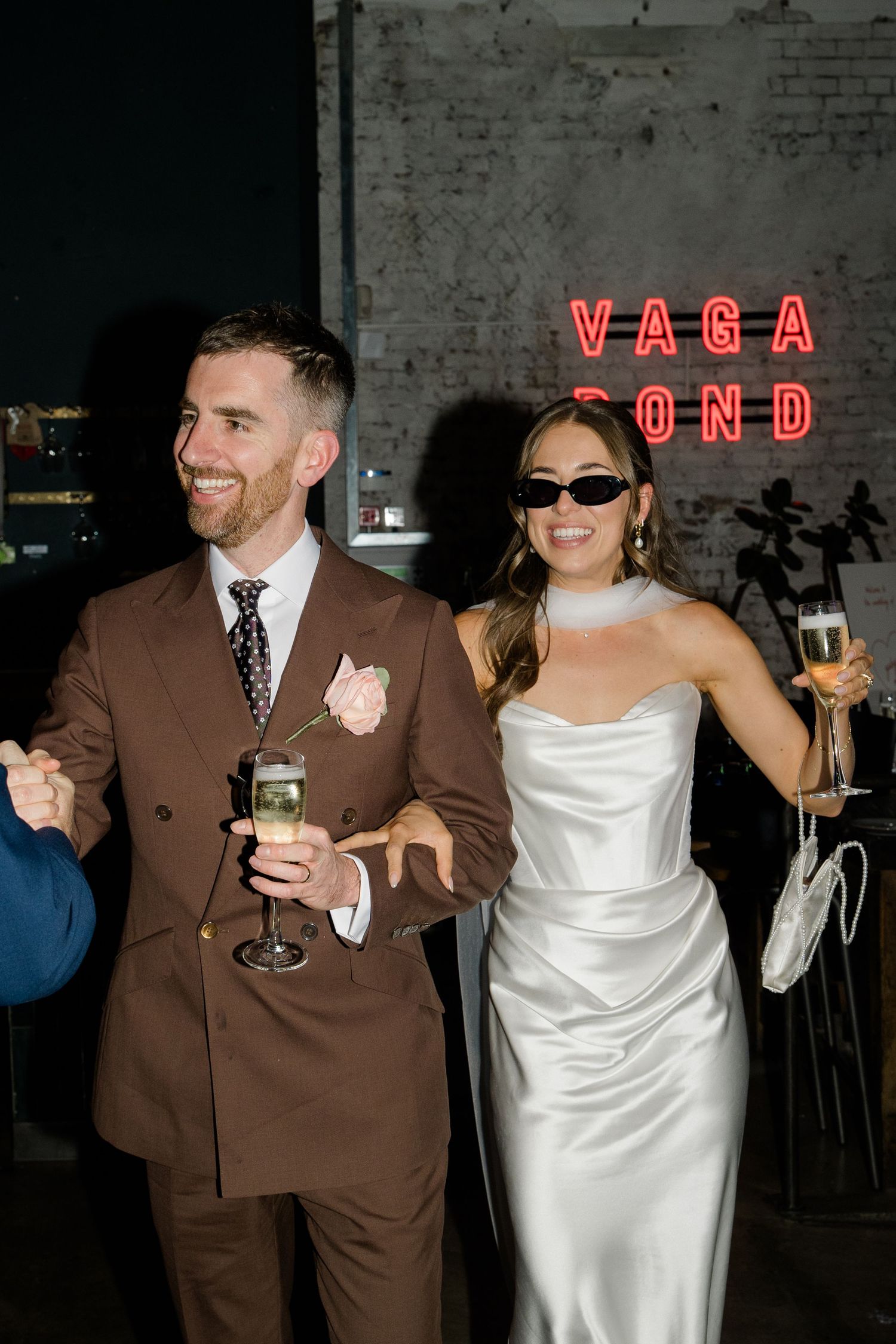 Smiling bride and groom in sunglasses holding champagne in front of a neon Vagabond sign.
