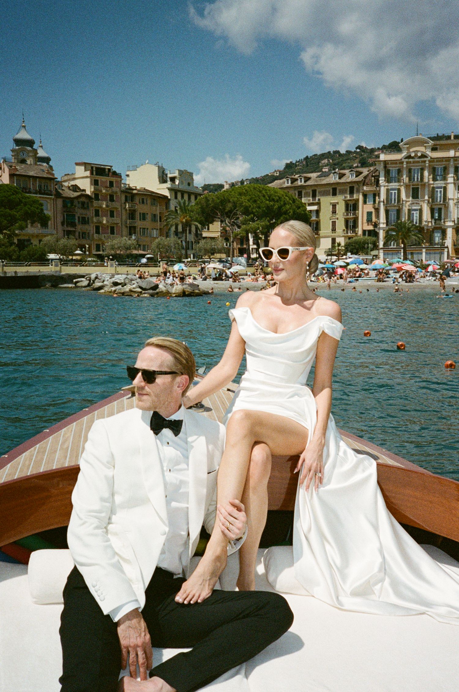 Bride and groom in white wedding attire pose on wooden boat with colorful Italian coastal town backdrop.