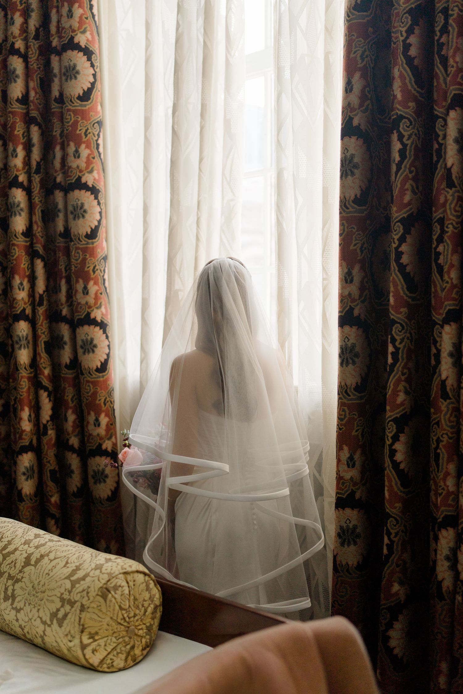 Bride in white wedding gown and veil sits by a window with ornate floral curtains on her wedding day.