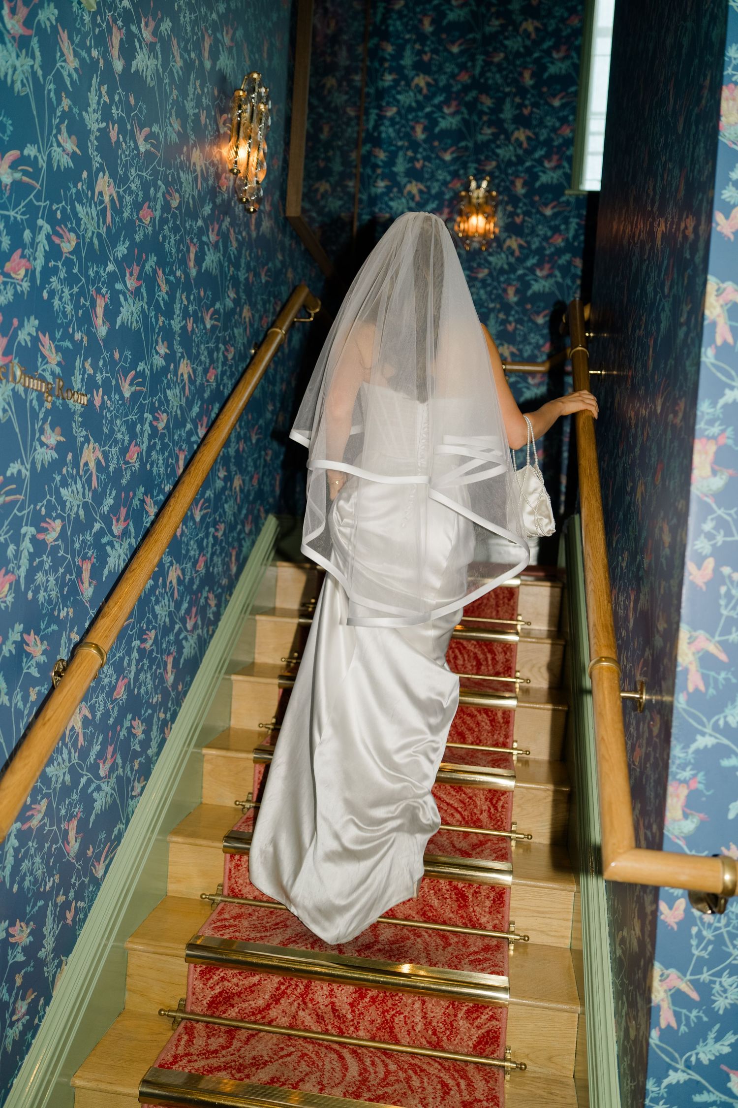 Bride in white wedding gown and veil ascending elegant staircase with red carpet and blue floral wallpaper.