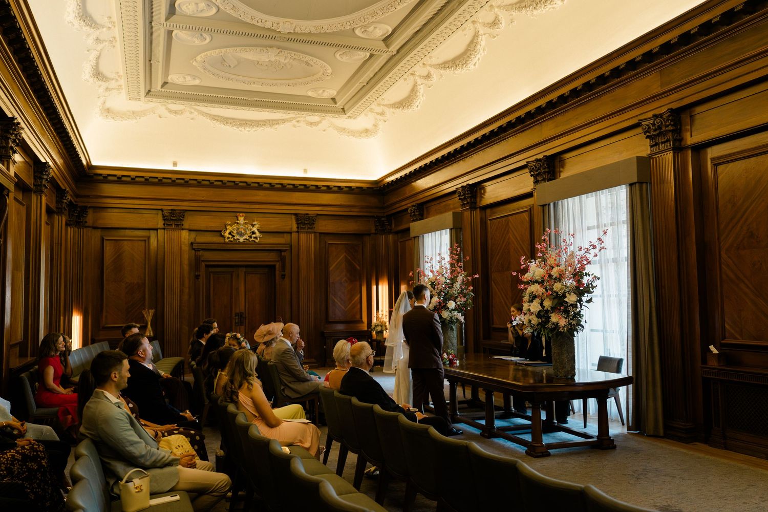 Wedding ceremony in elegant wood-paneled room with ornate ceiling, guests seated facing couple at altar.