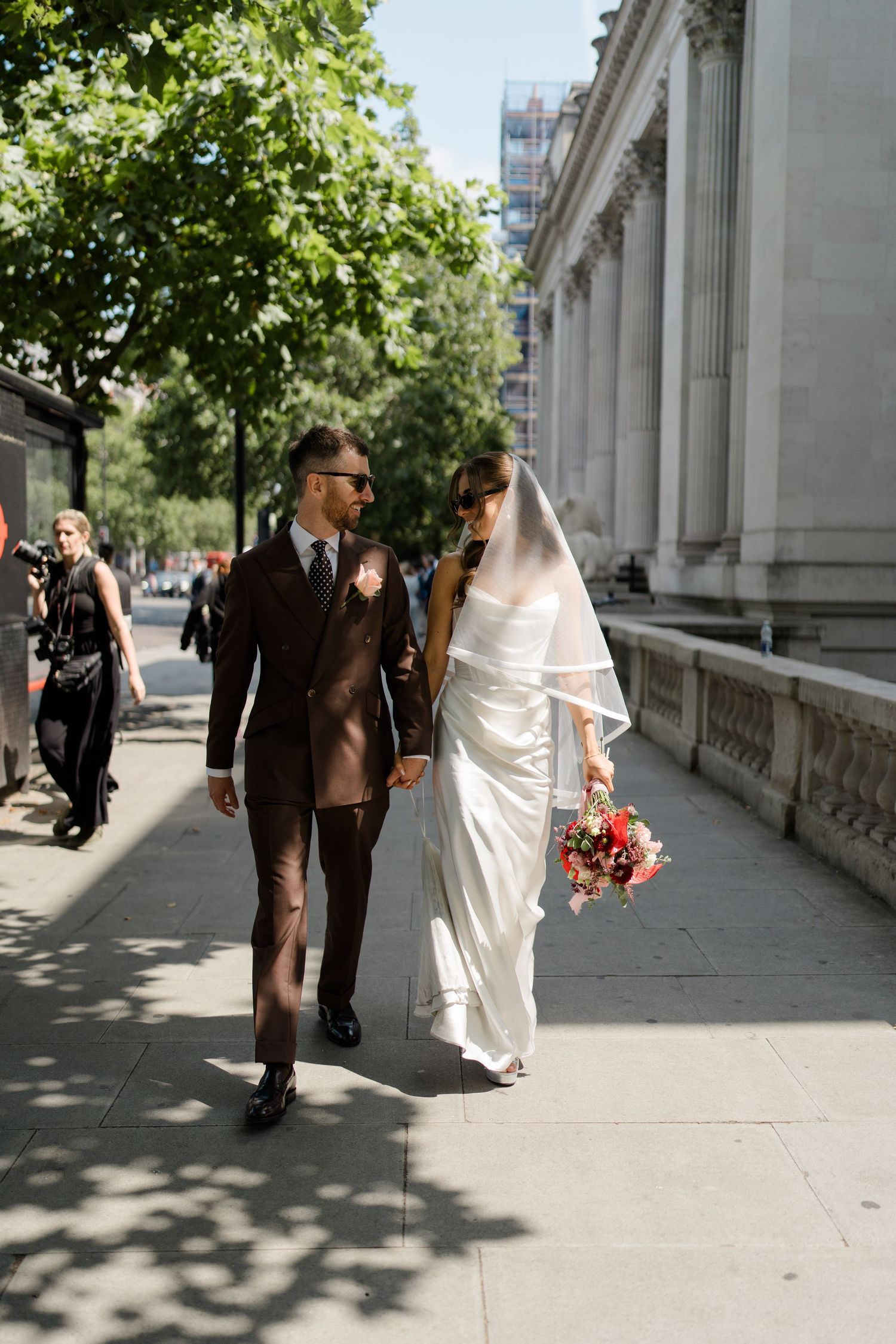 Bride and groom walking hand in hand along sunny city sidewalk beside classical stone building, bride wearing flowing veil.