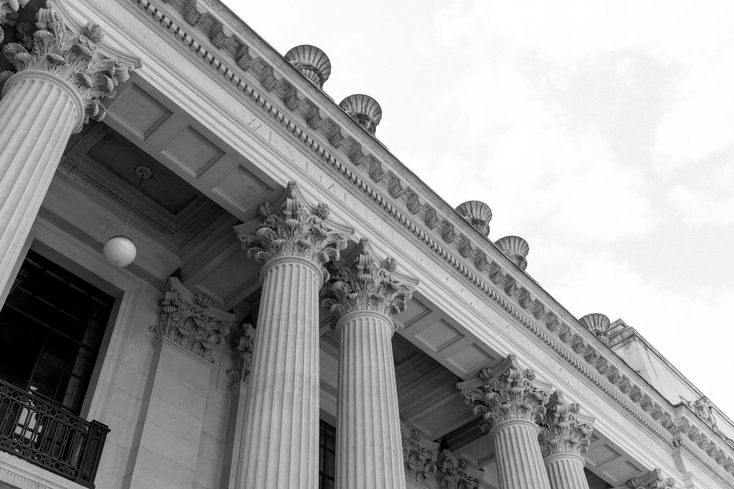 Black and white photo of neoclassical building facade with ornate Corinthian columns against cloudy sky.
