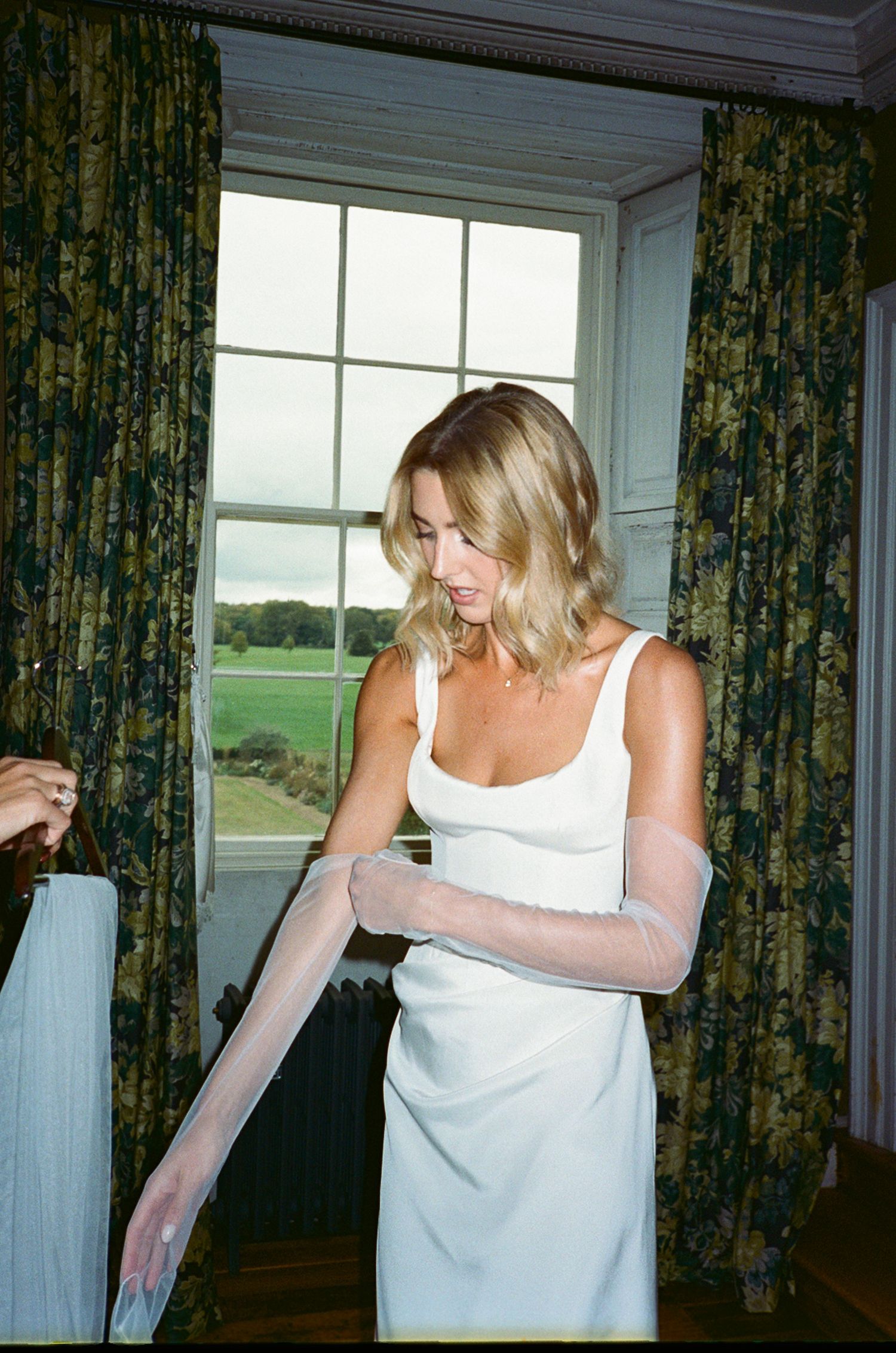 Blonde bride in sleek white gown putting on sheer gloves by a large window with garden views.
