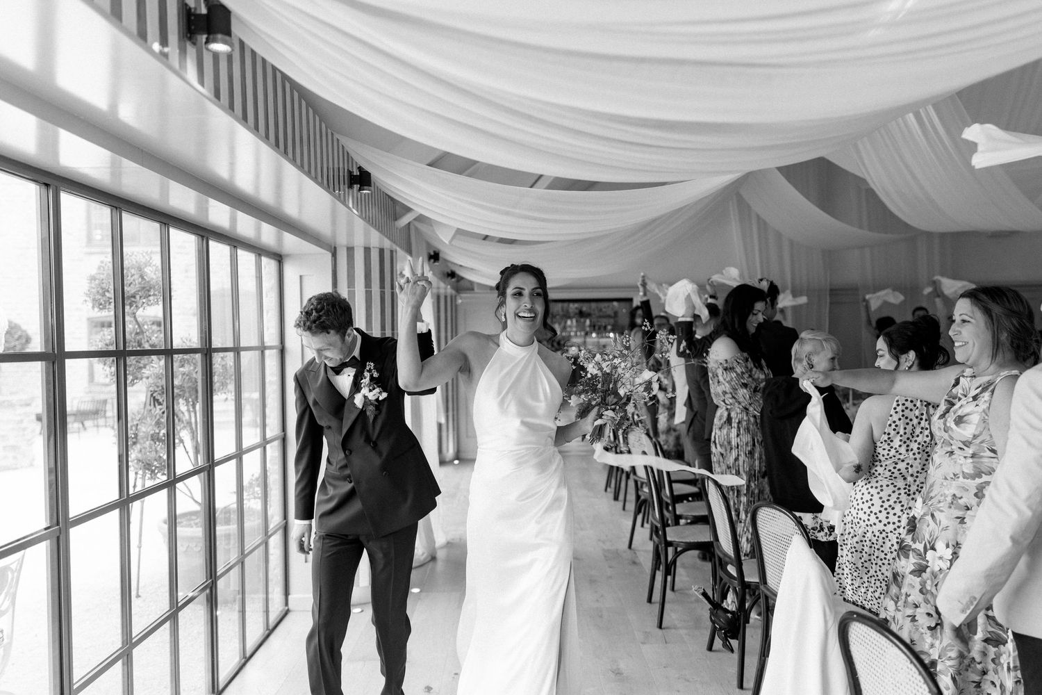 Black and white photo of bride and groom entering wedding reception tent to applause from guests.