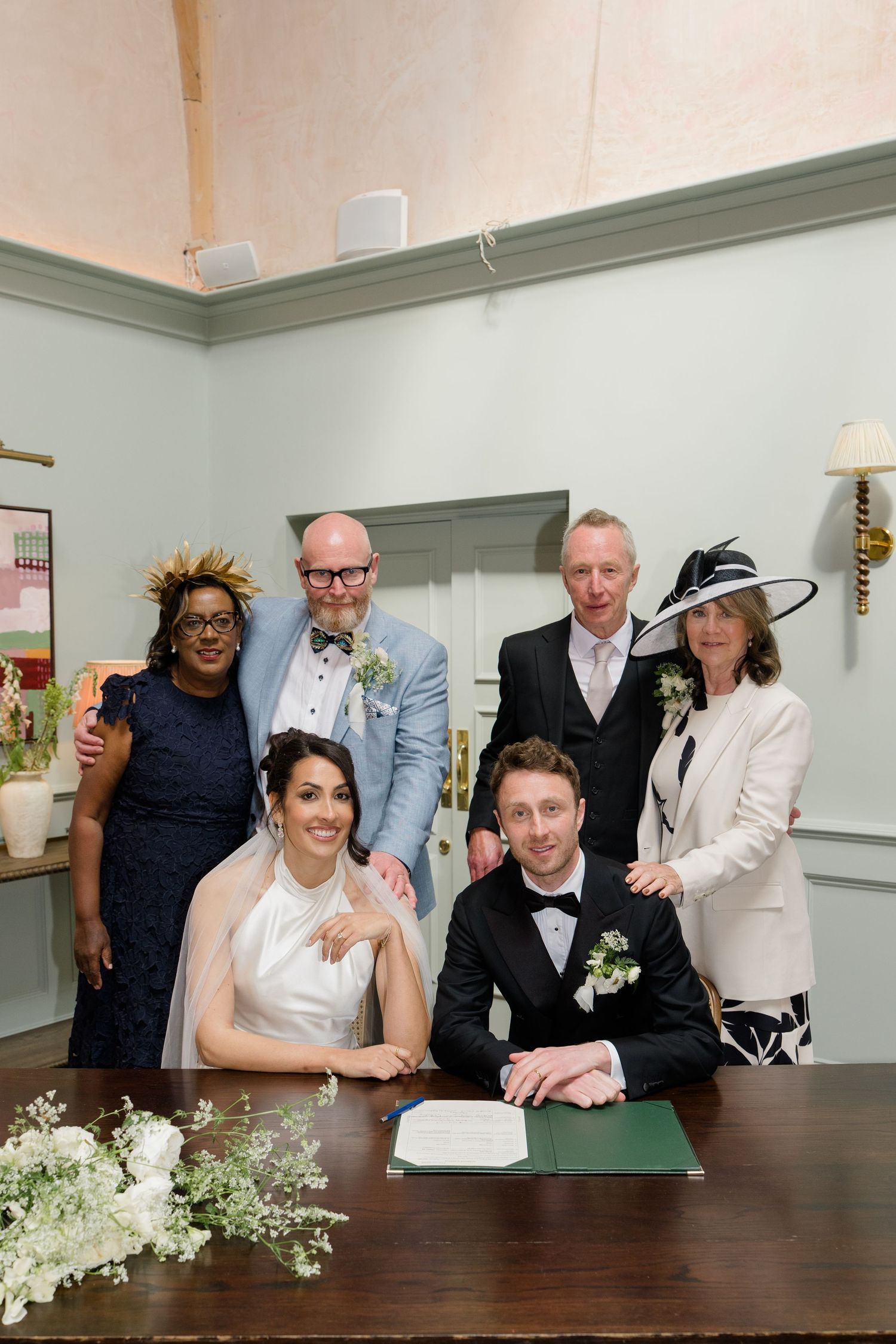 Bride and groom sign wedding register surrounded by family witnesses in a elegant pale green room.