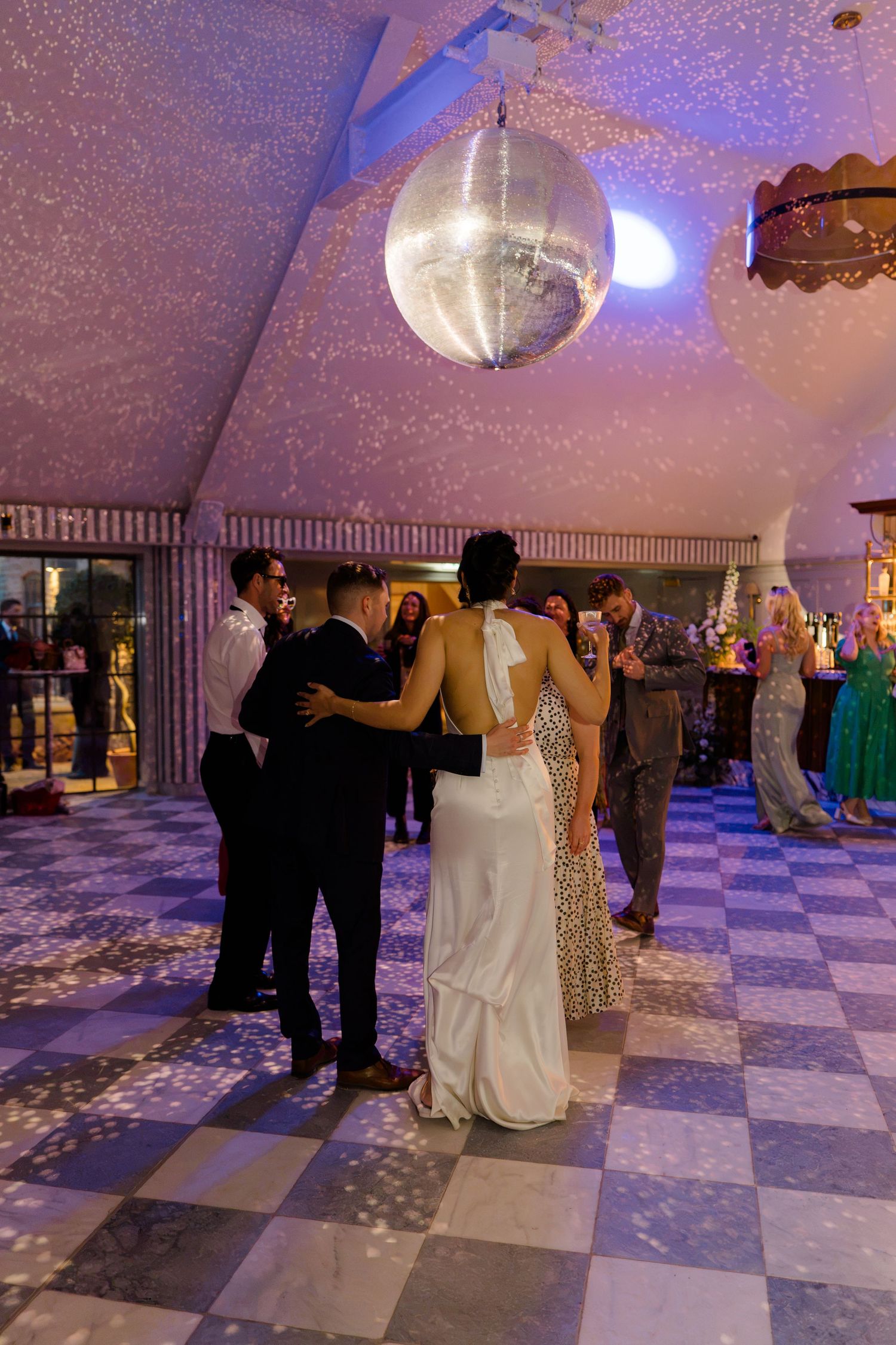 Bride and groom share first dance under glittering disco ball on checkered floor at wedding reception.