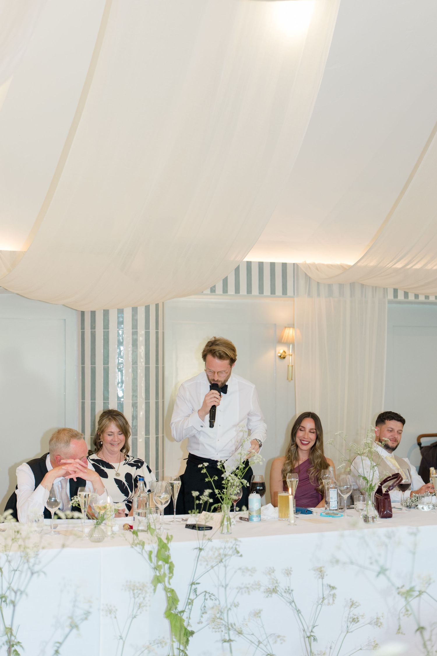 Best man delivering speech at wedding reception while guests laugh at head table.