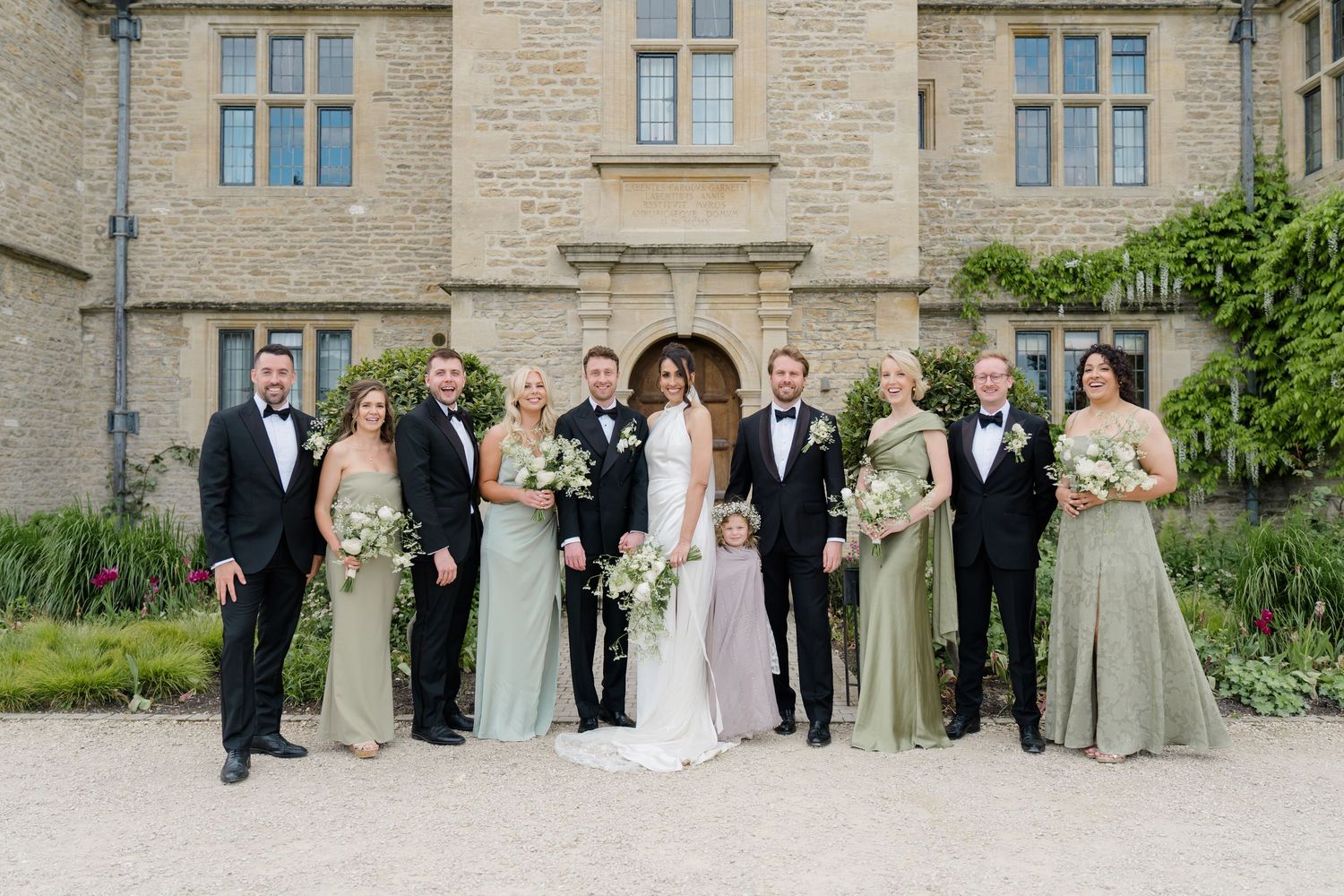 Wedding party poses in front of stone manor, bridesmaids in sage green gowns and groomsmen in tuxedos.