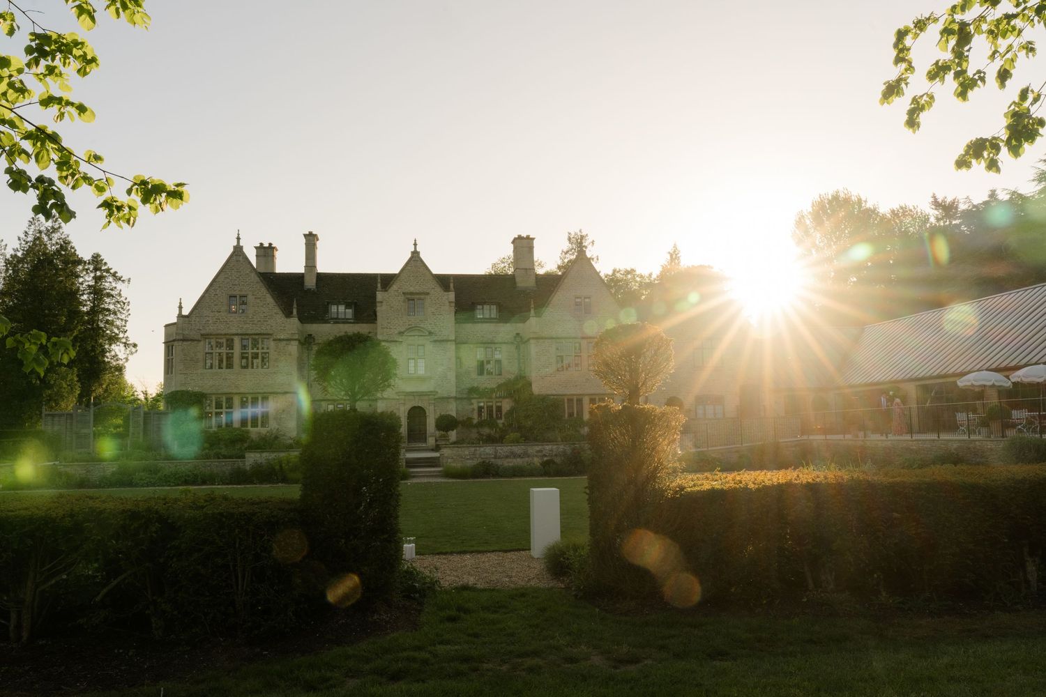 Golden sunset bursting through trees behind an elegant stone English country manor house and garden.