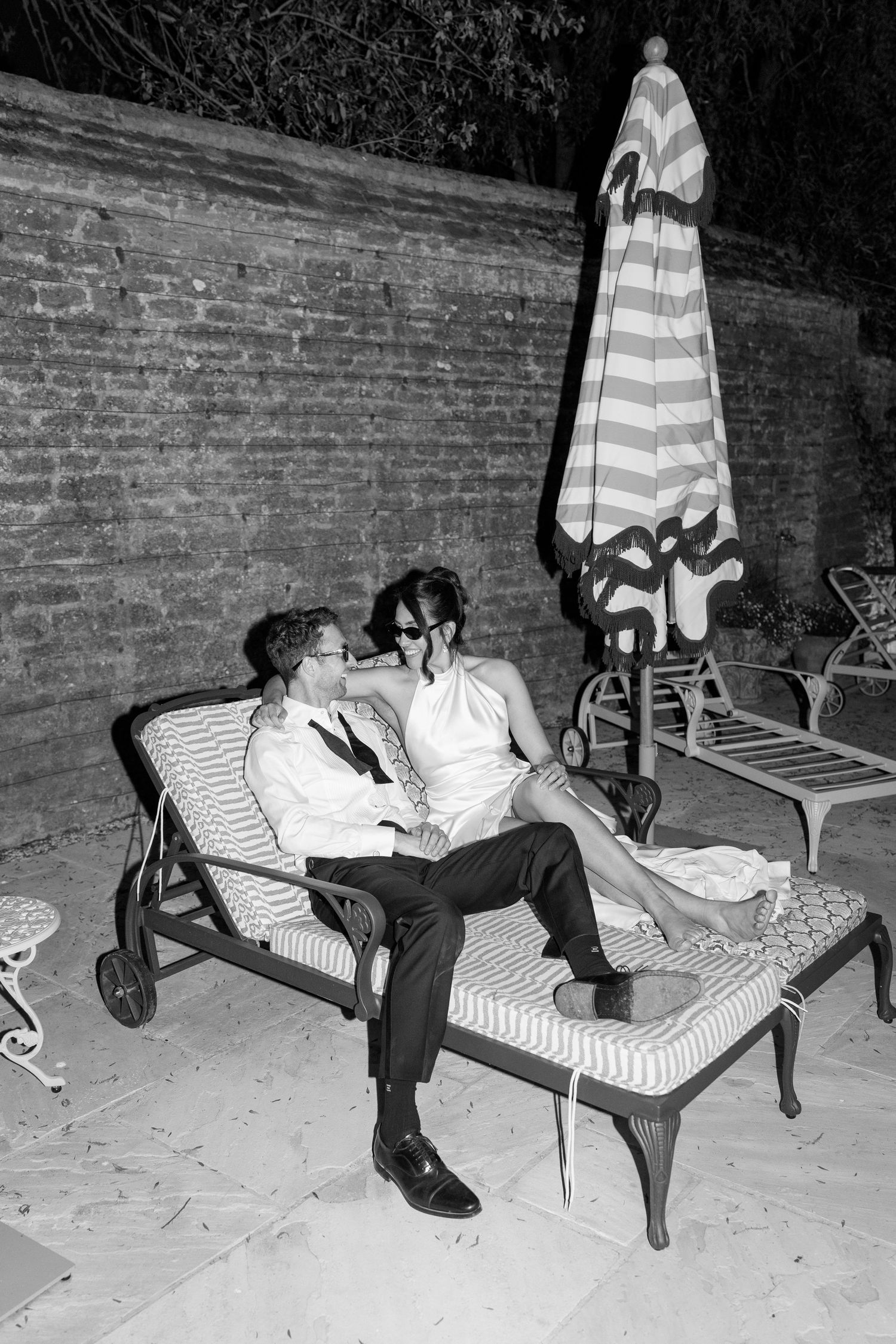 Bride and groom relax together on lounge chairs by brick wall at night after wedding celebration.